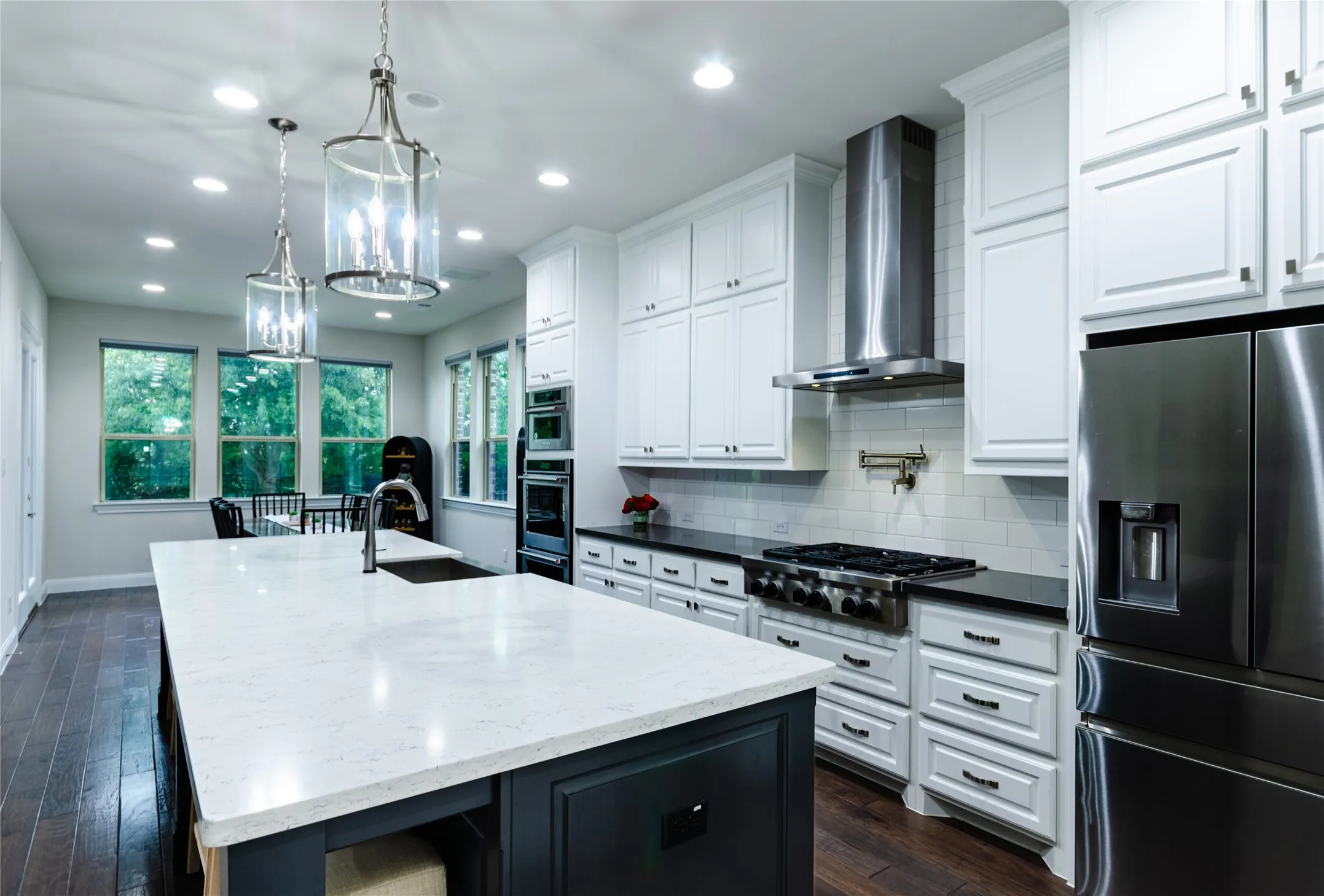 Gourmet kitchen featuring a large island with a light-toned countertop and integrated sink, white shaker-style cabinetry, dark wood-finish flooring, stainless steel appliances, and dual pendant lighting fixtures