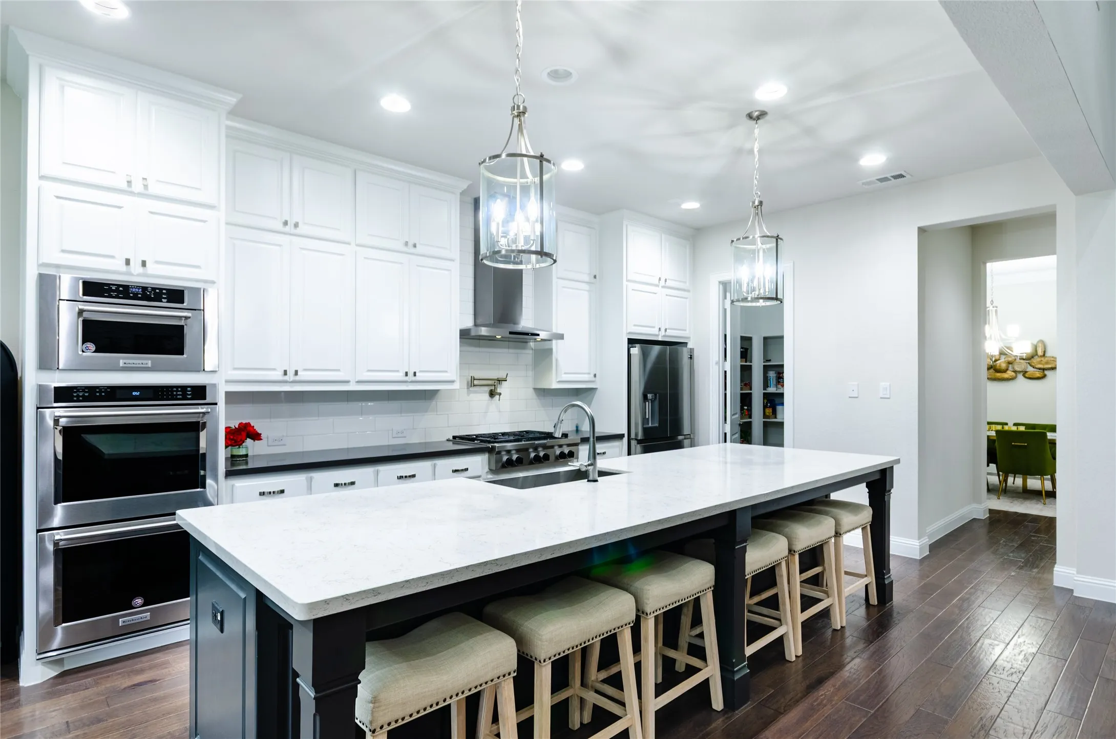 Gourmet kitchen featuring a large central island with seating, white shaker cabinetry, stainless steel appliances including double ovens and a range hood, and dark wood-finish flooring