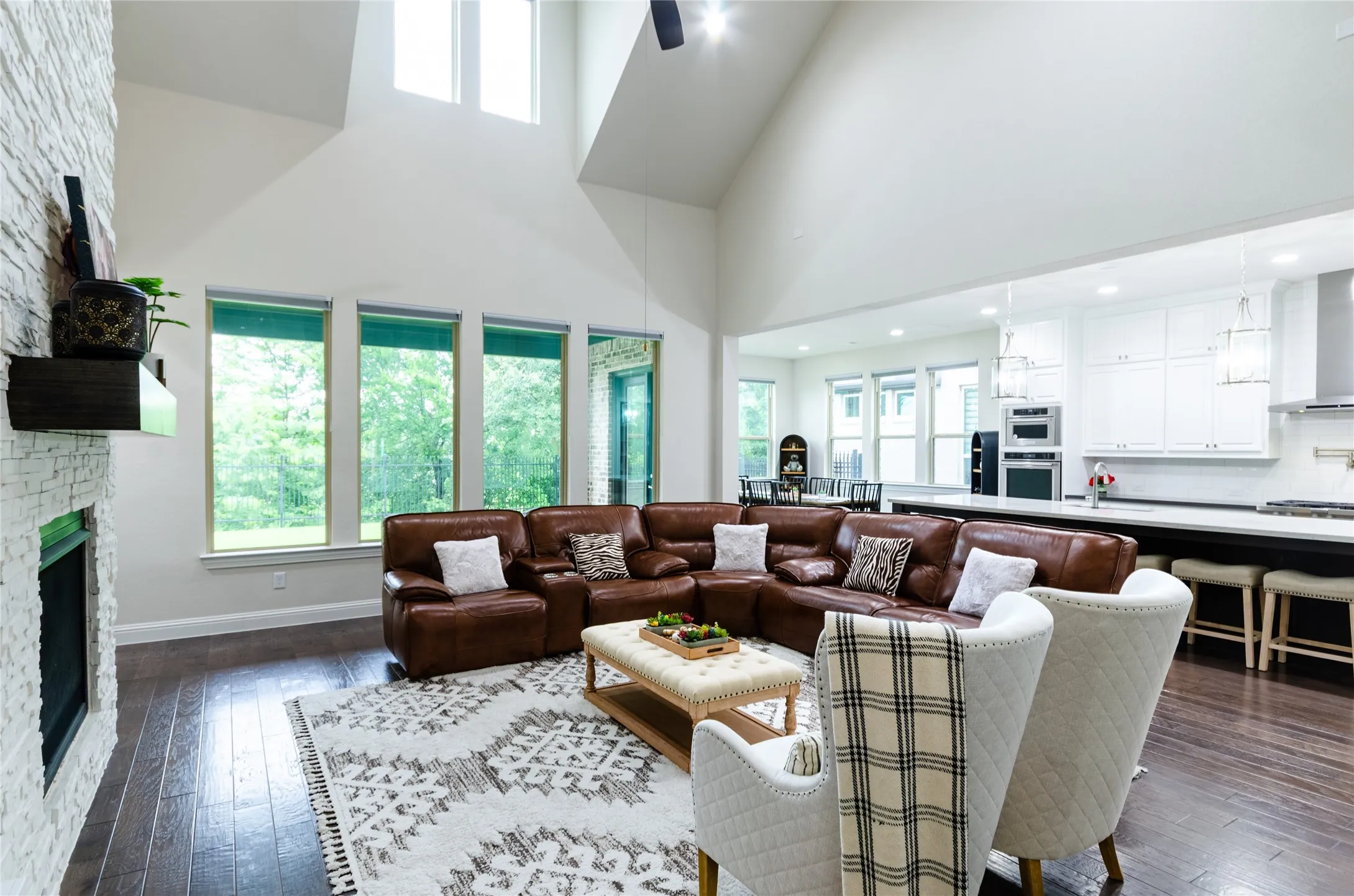 Open-concept living space featuring high vaulted ceilings, expansive windows, a stacked stone fireplace, wood-finish flooring, and a kitchen with white cabinetry and stainless steel appliances