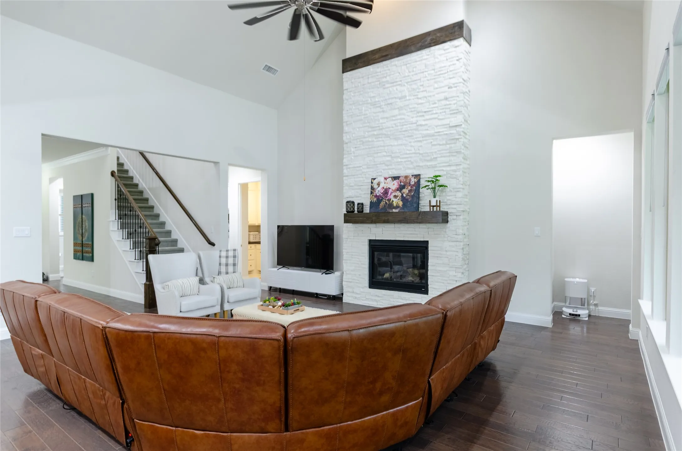 Two-story living area featuring a stacked stone fireplace with a substantial wood mantel, dark wood-finish flooring, and a ceiling fan
