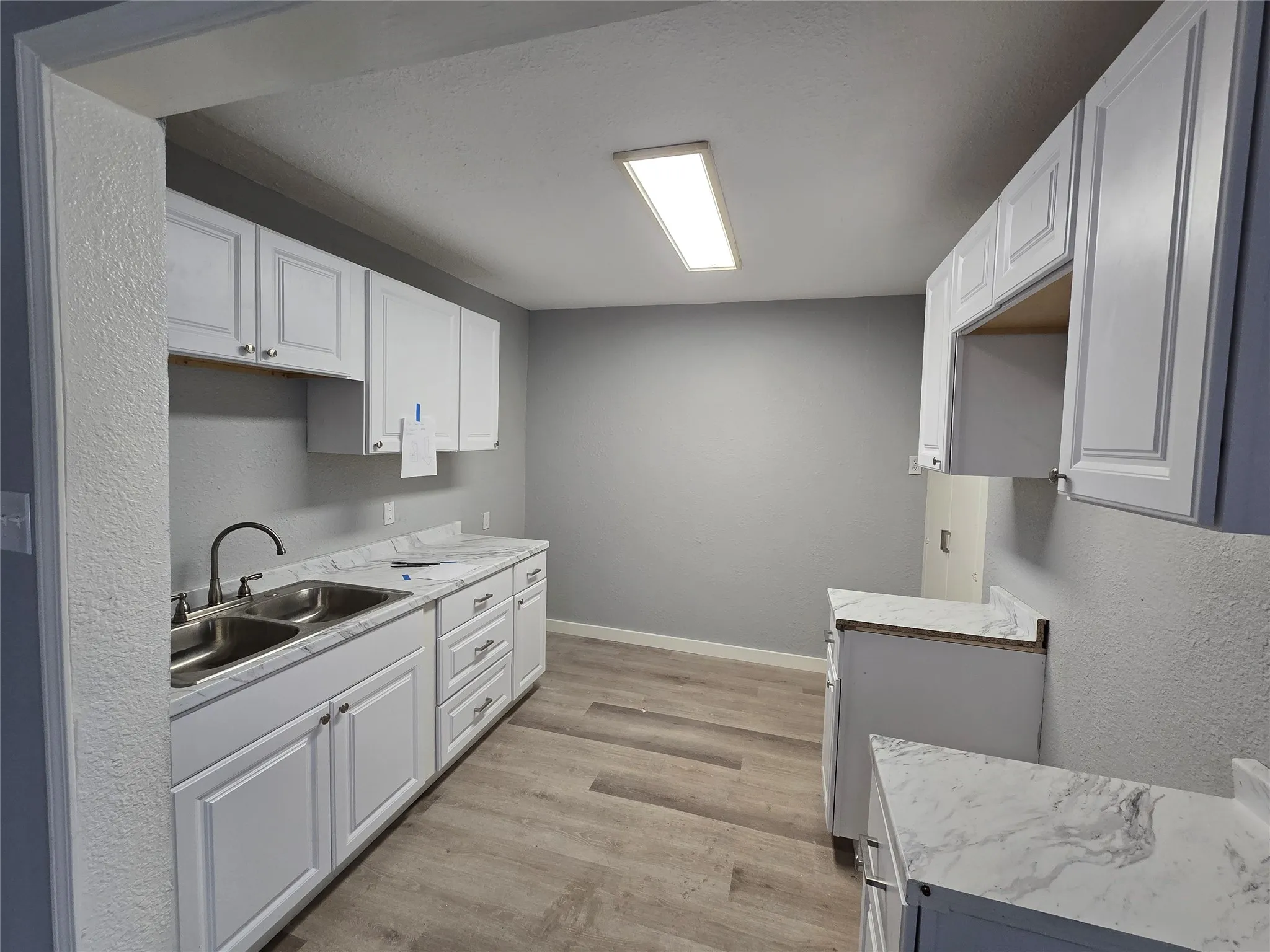 White cabinetry with chrome hardware, dual basin stainless steel sink, black gooseneck faucet, light-toned wood-finish flooring, and white countertops