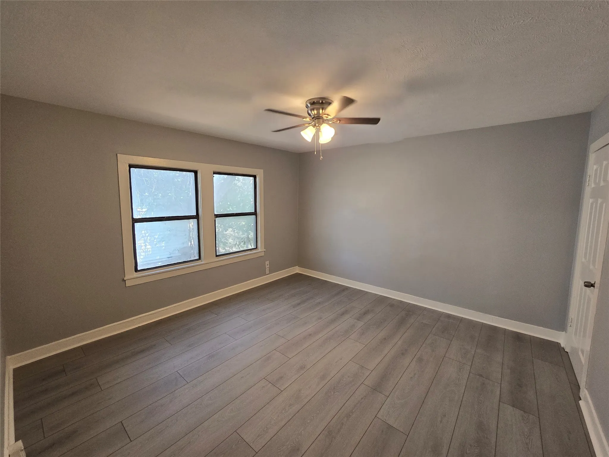 Wood-finish flooring, two large windows with white trim, a ceiling fan with integrated lighting, and a white panel door