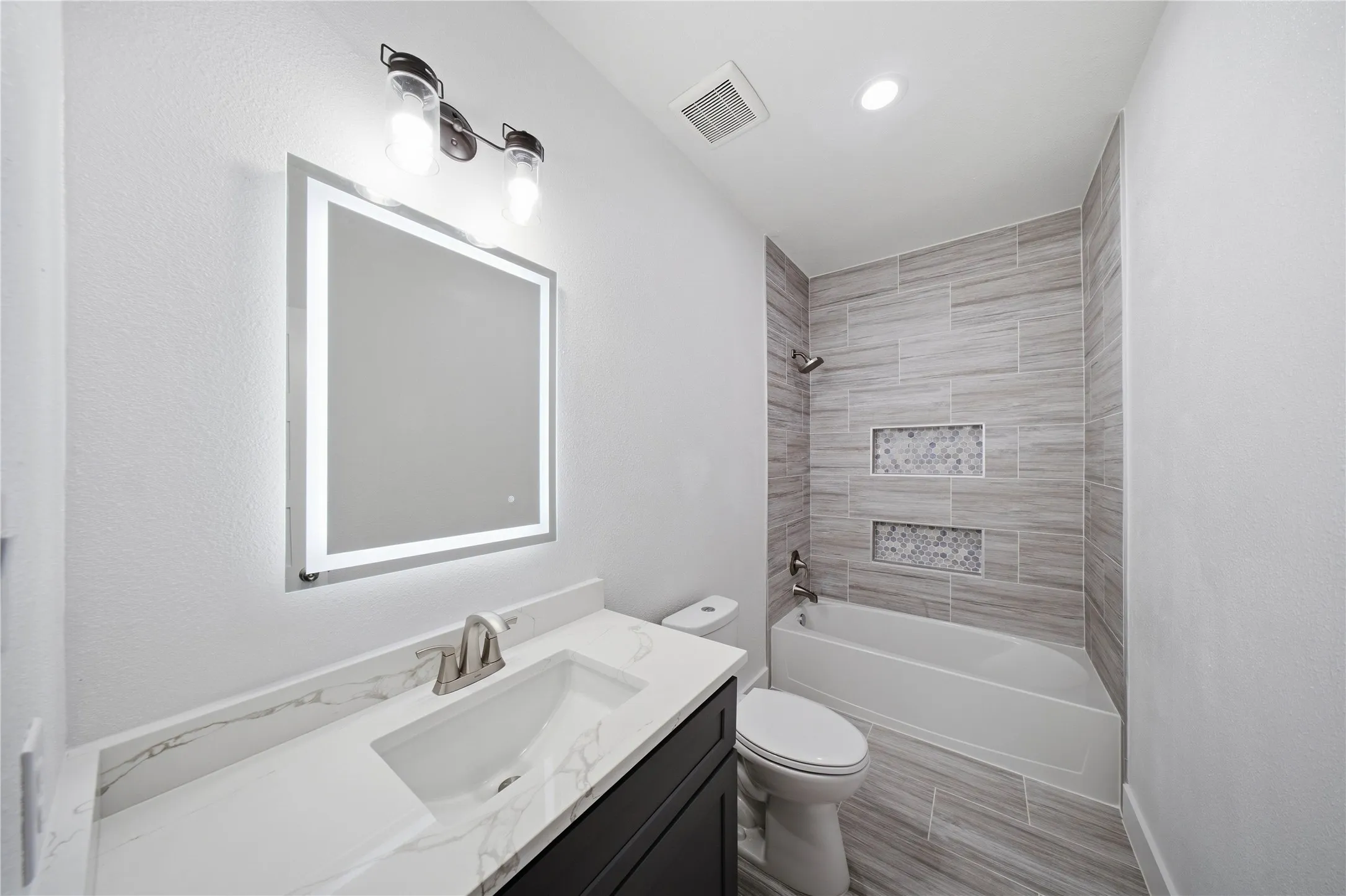 Vanity with a rectangular undermount sink, a countertop with veining, an illuminated mirror, a tile-surround bathtub with built-in niches, and wood-finish tile flooring