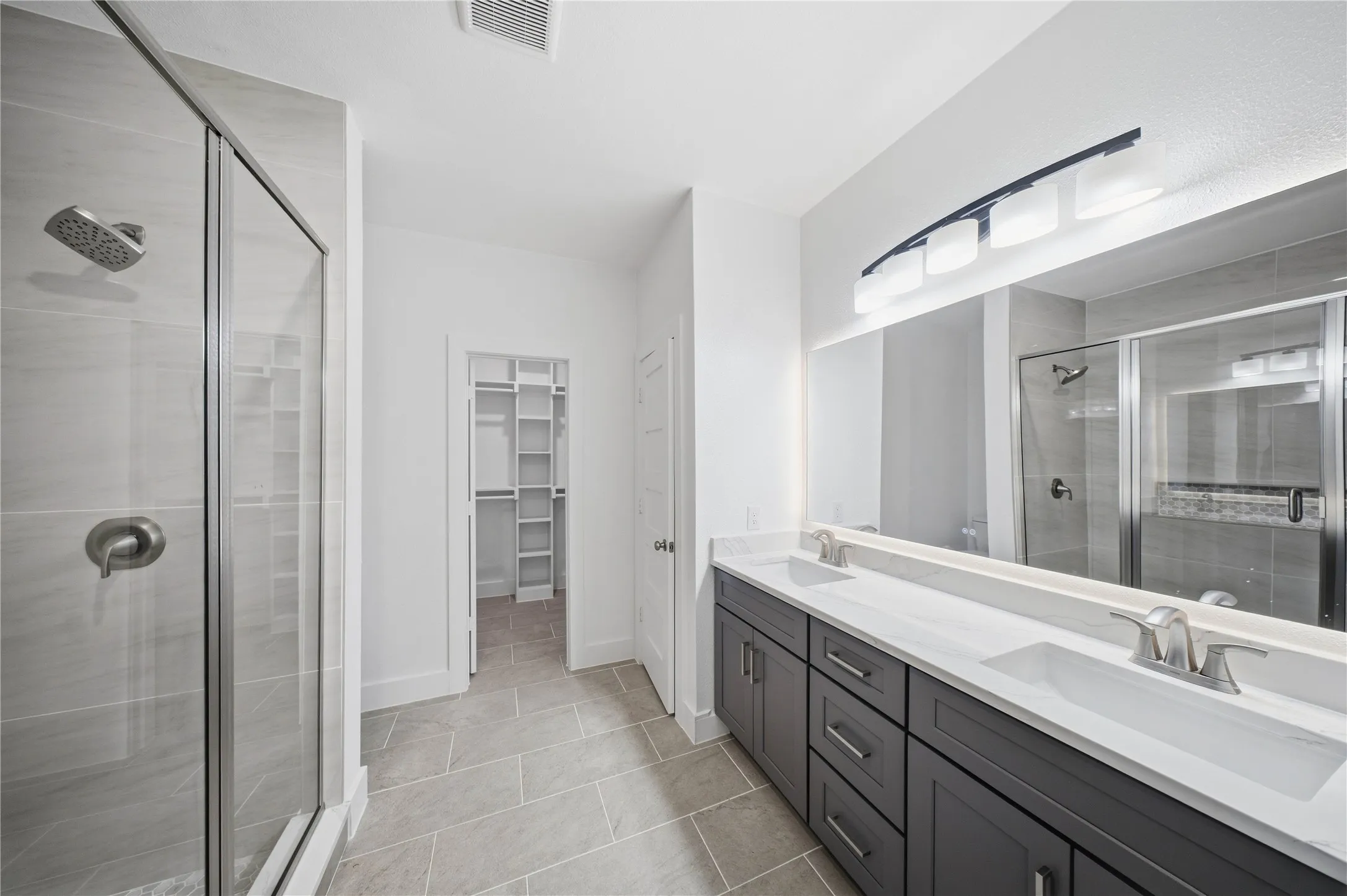 Double vanity with undermount sinks and dark cabinetry, large mirror, glass-enclosed shower with tile surround, and walk-in closet with shelving