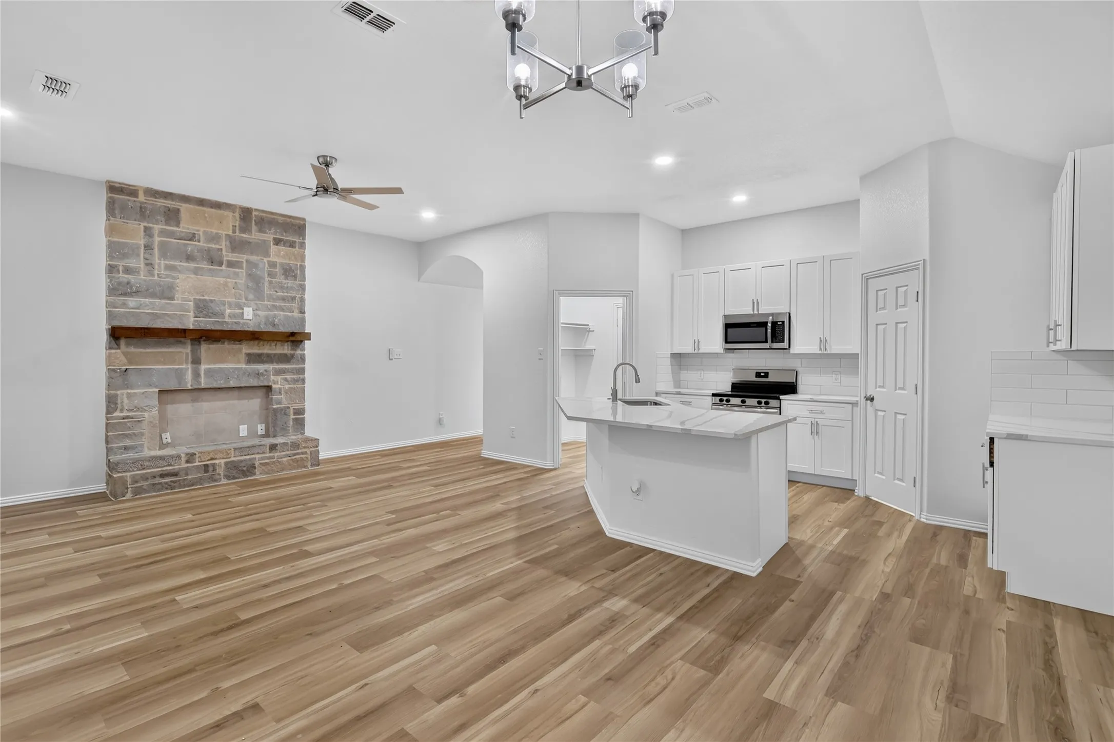 Wood-finish flooring, stone fireplace with wood mantel, white cabinetry, island with sink, and subway tile backsplash