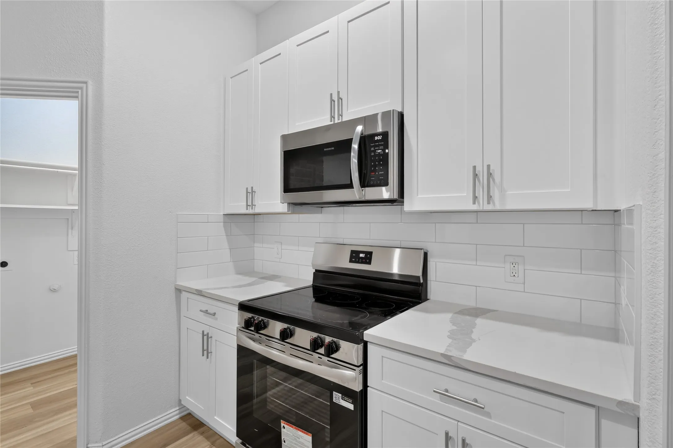 White shaker-style cabinetry with brushed nickel hardware, stainless steel appliances, white subway tile backsplash, light-toned countertops, and wood-finish flooring