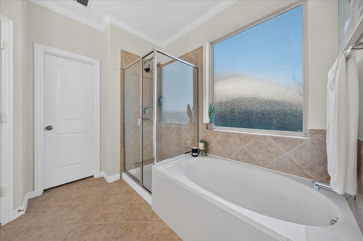Built-in soaking tub, glass-enclosed shower, large privacy window, and neutral-toned floor tiling
