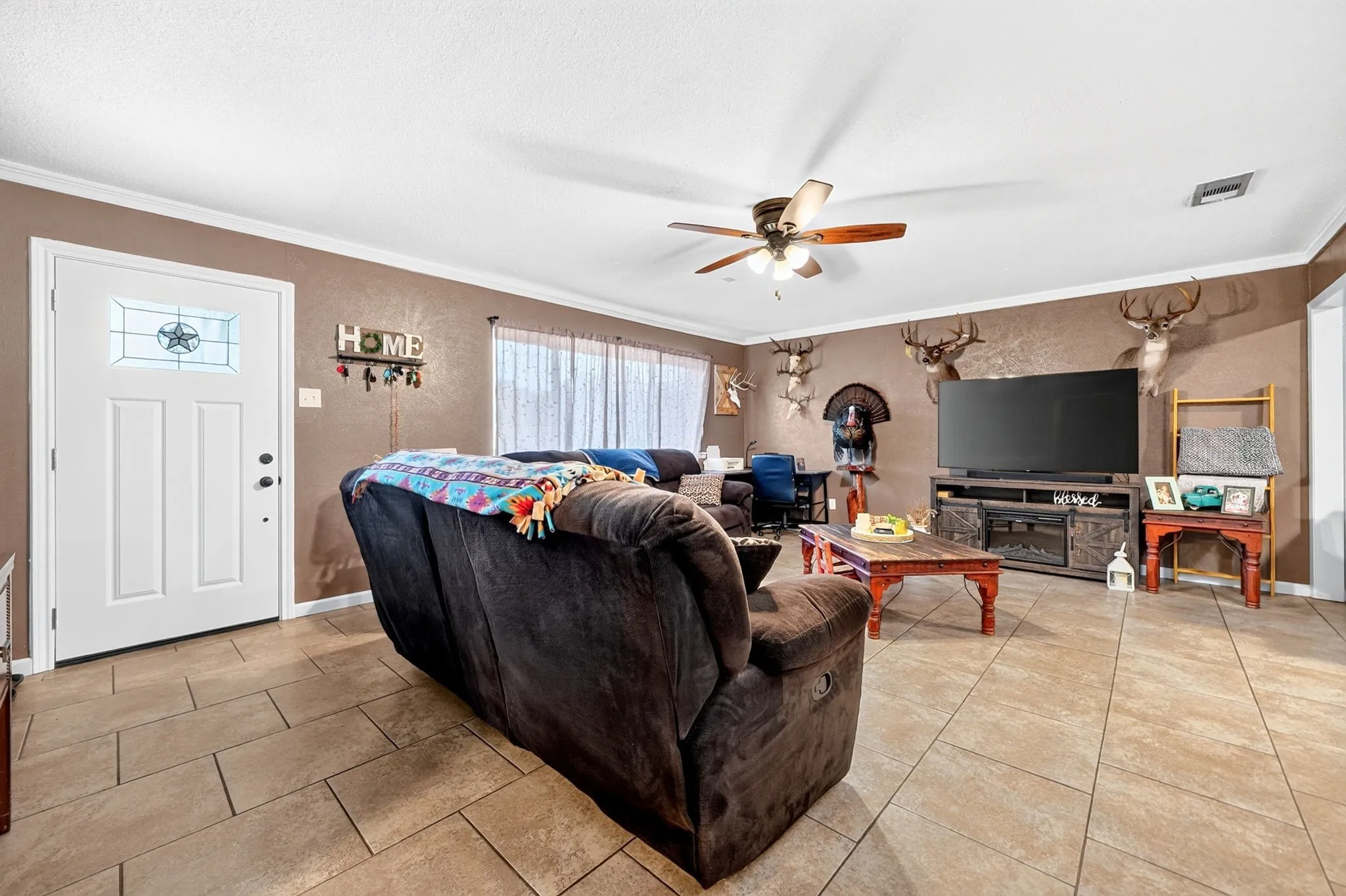 Living area featuring tile flooring, a white paneled entry door with decorative glass, crown molding, and a ceiling fan with light fixture