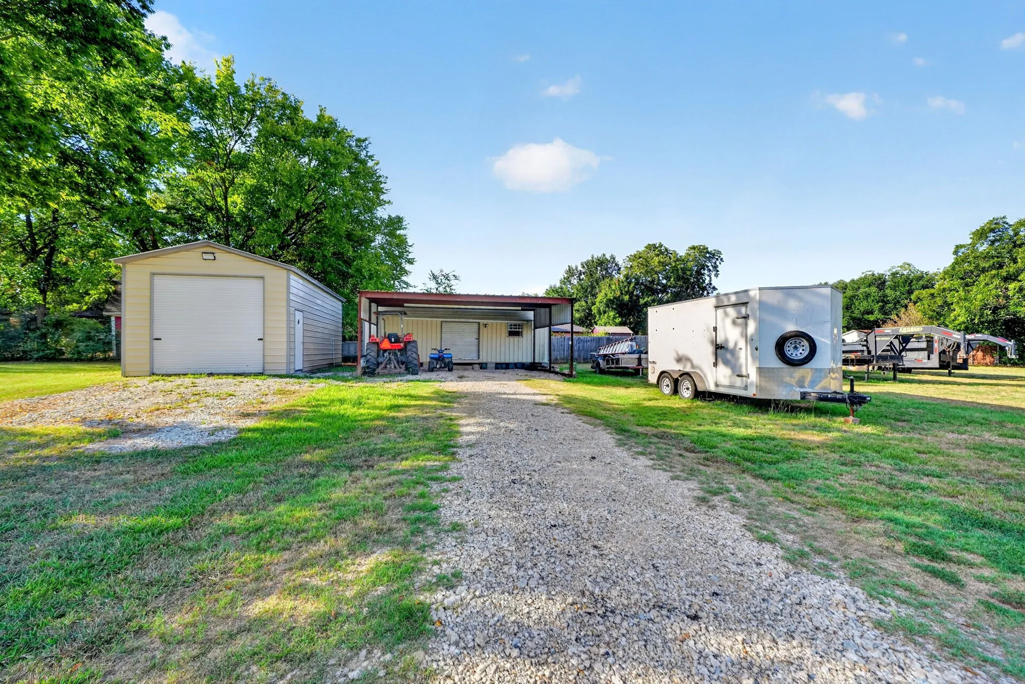 Gravel driveway leading to a metal garage with a roll-up door, an open-sided metal structure, and a secondary building
