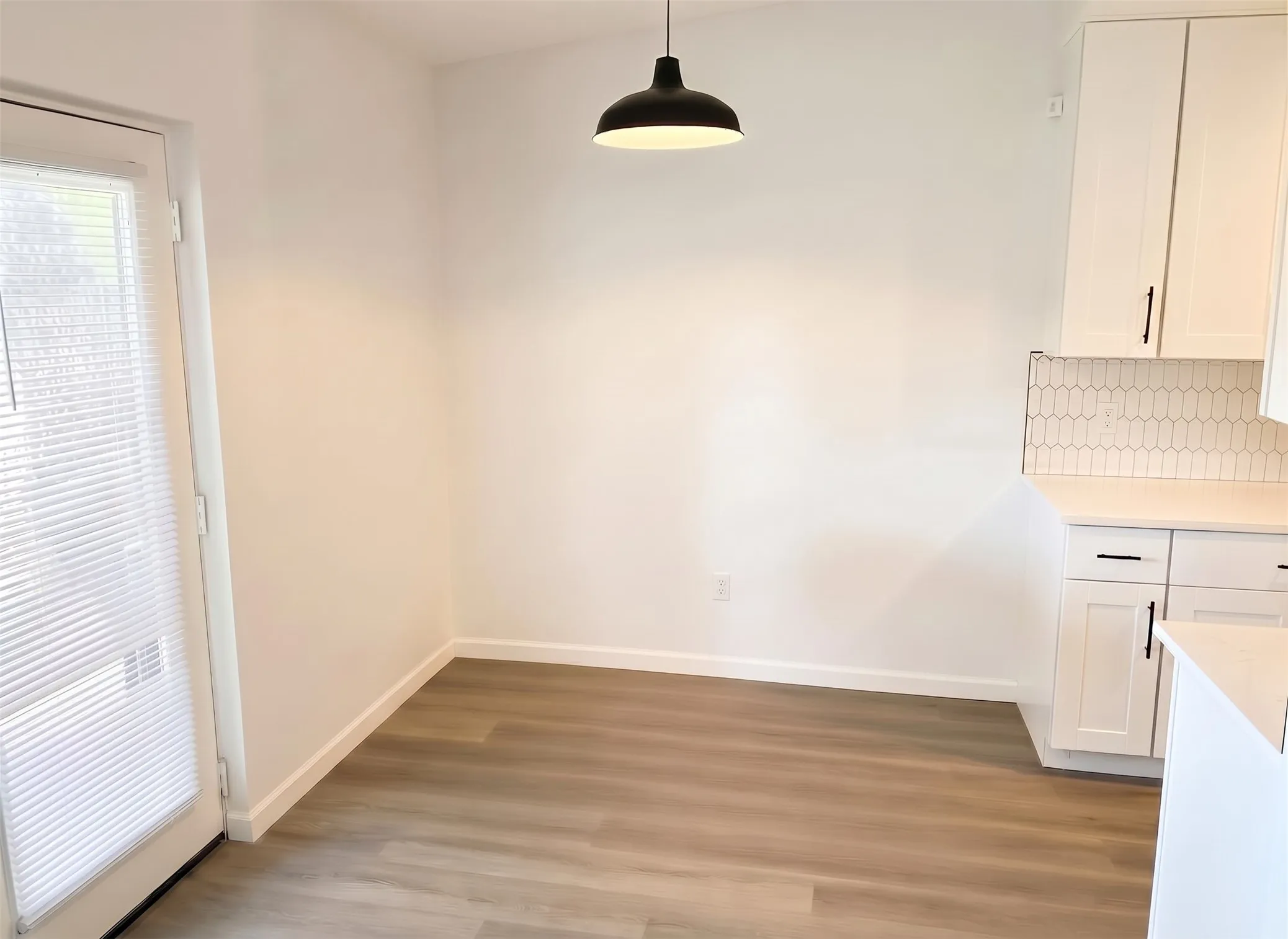 Wood-finish flooring, white cabinetry with dark hardware, white countertop, white picket tile backsplash, and a modern black pendant light fixture