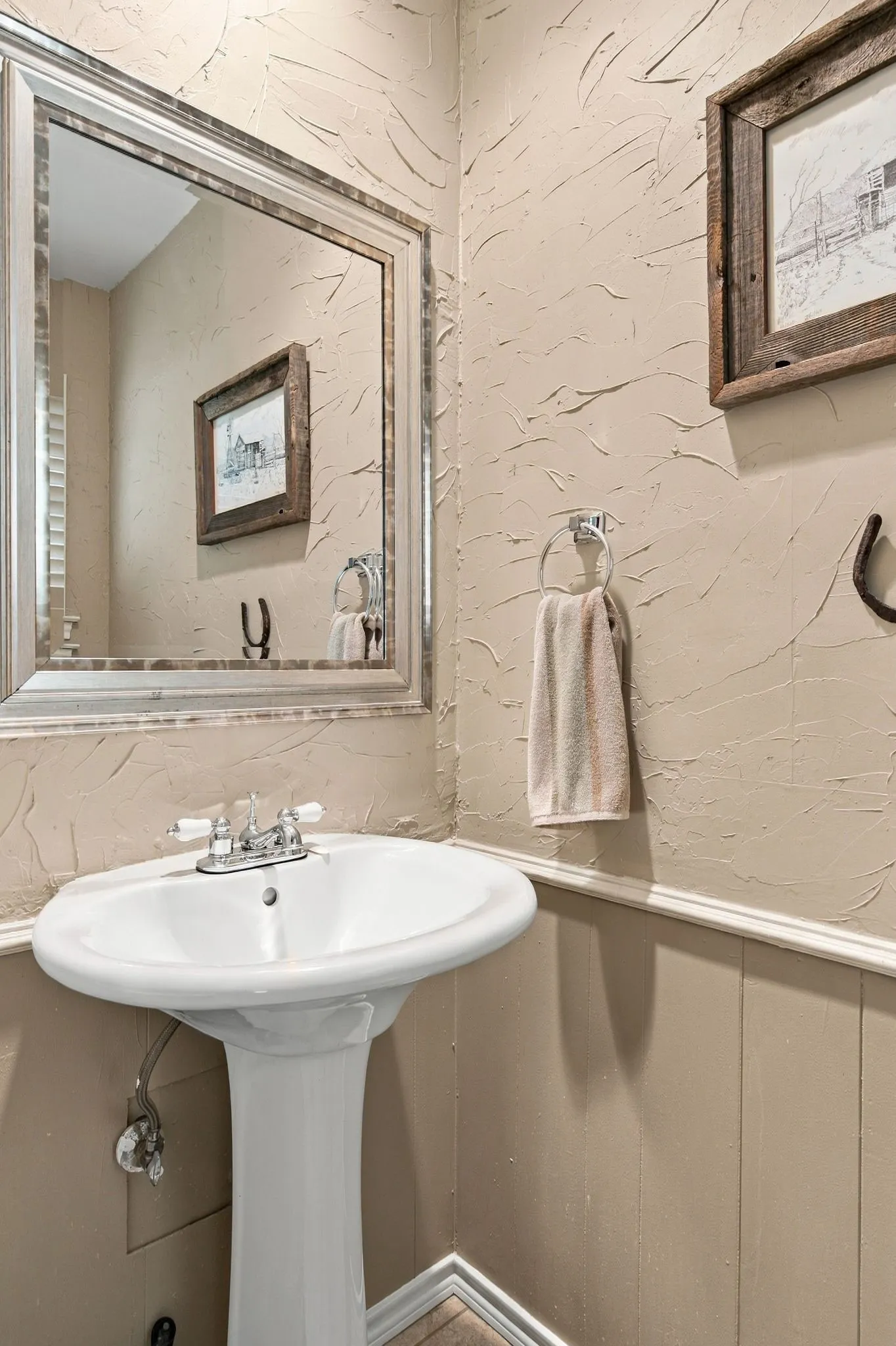 Pedestal sink with chrome faucet, textured wall finish, wainscoting, and decorative framed mirror