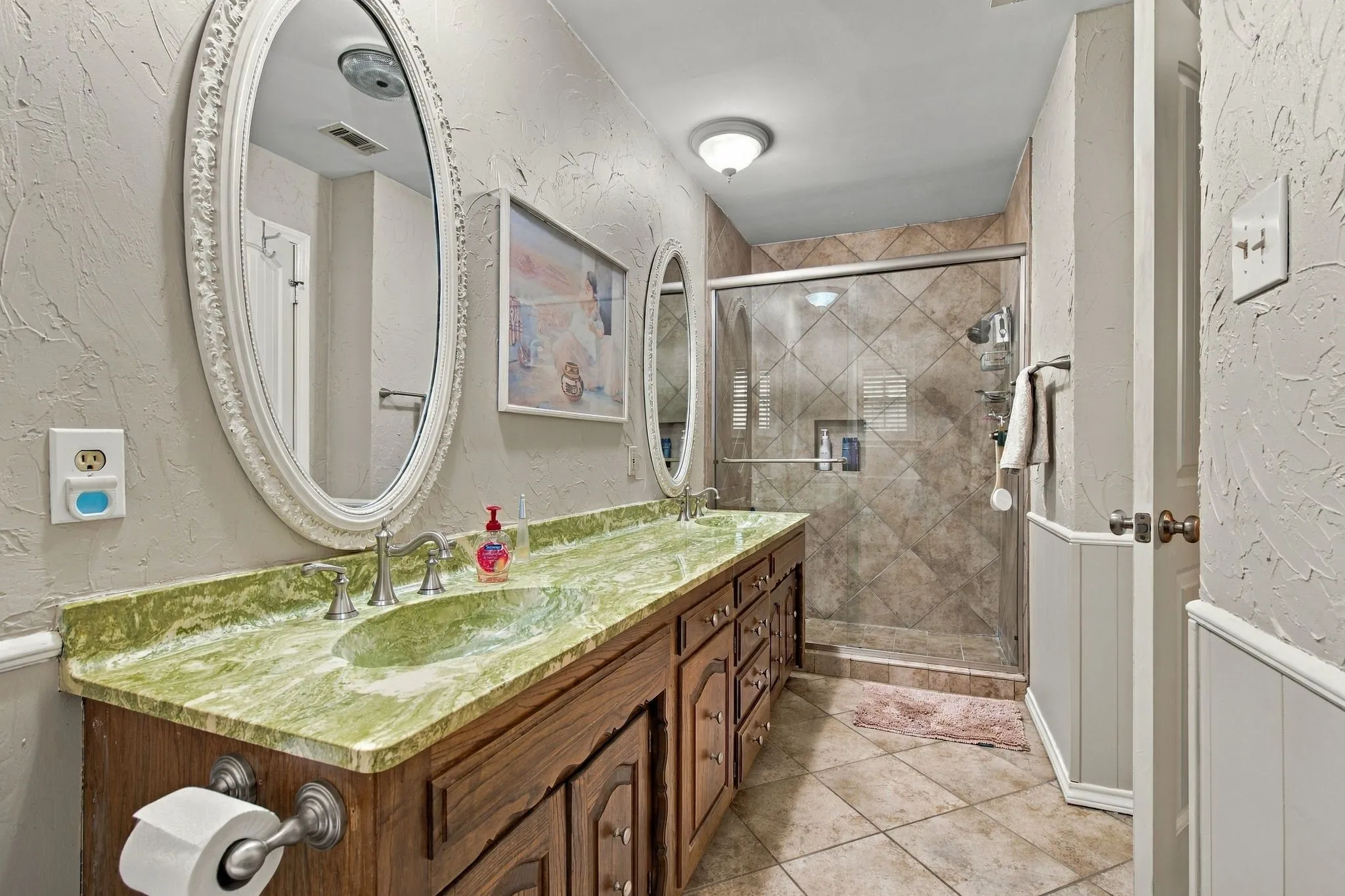 Double vanity with green natural stone countertop, wood-finish cabinetry, and oval mirrors