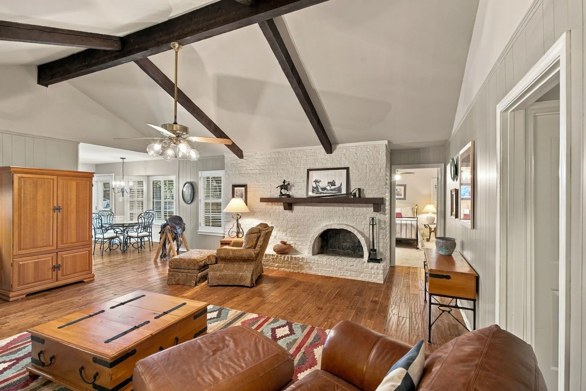 Vaulted ceilings with exposed beams, wood-finish flooring, white brick fireplace with mantel, ceiling fan with light fixture, and windows with white blinds
