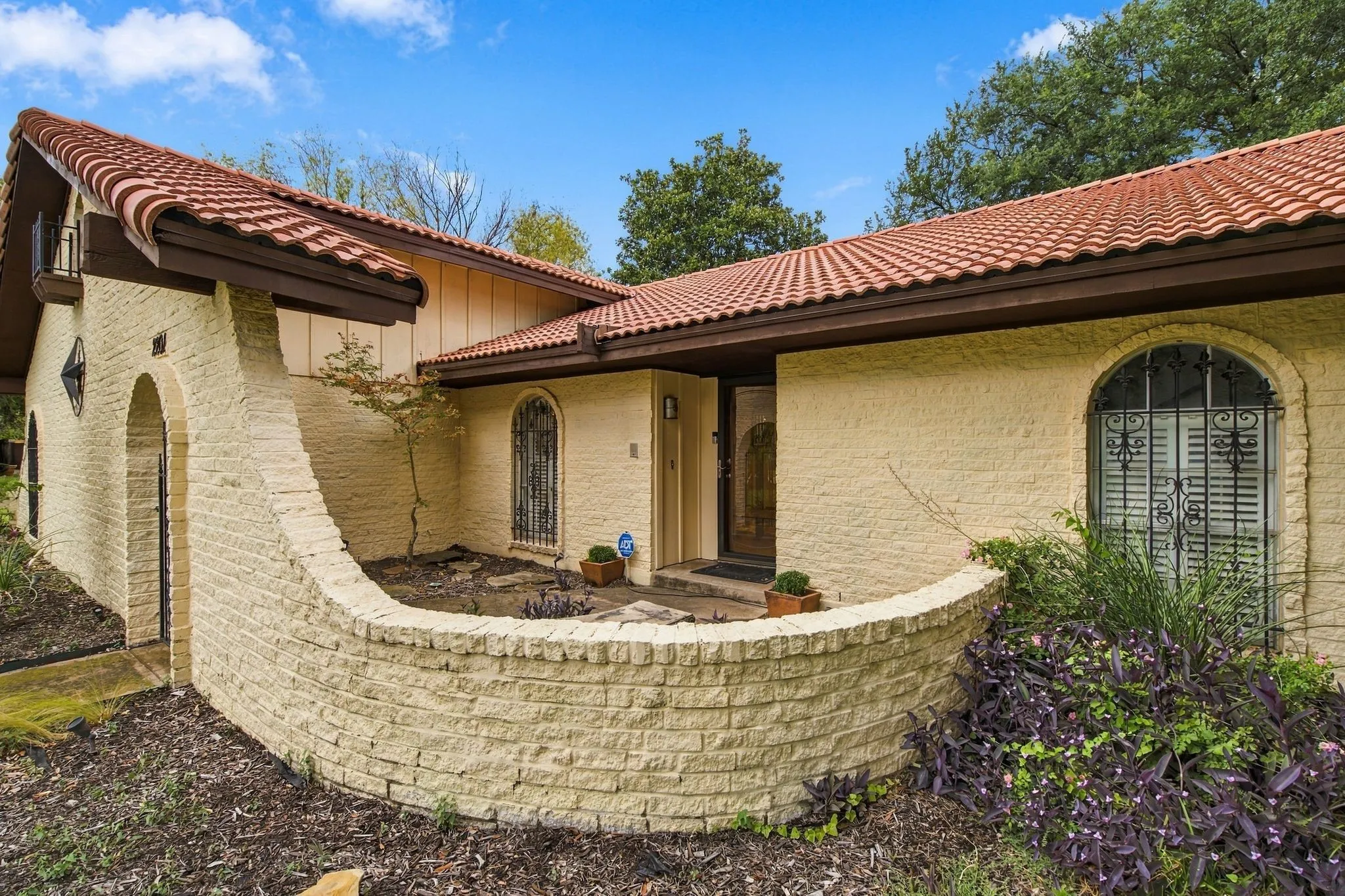 Terracotta tile roof, textured stucco exterior, curved stone retaining wall, arched entry, and decorative window grilles