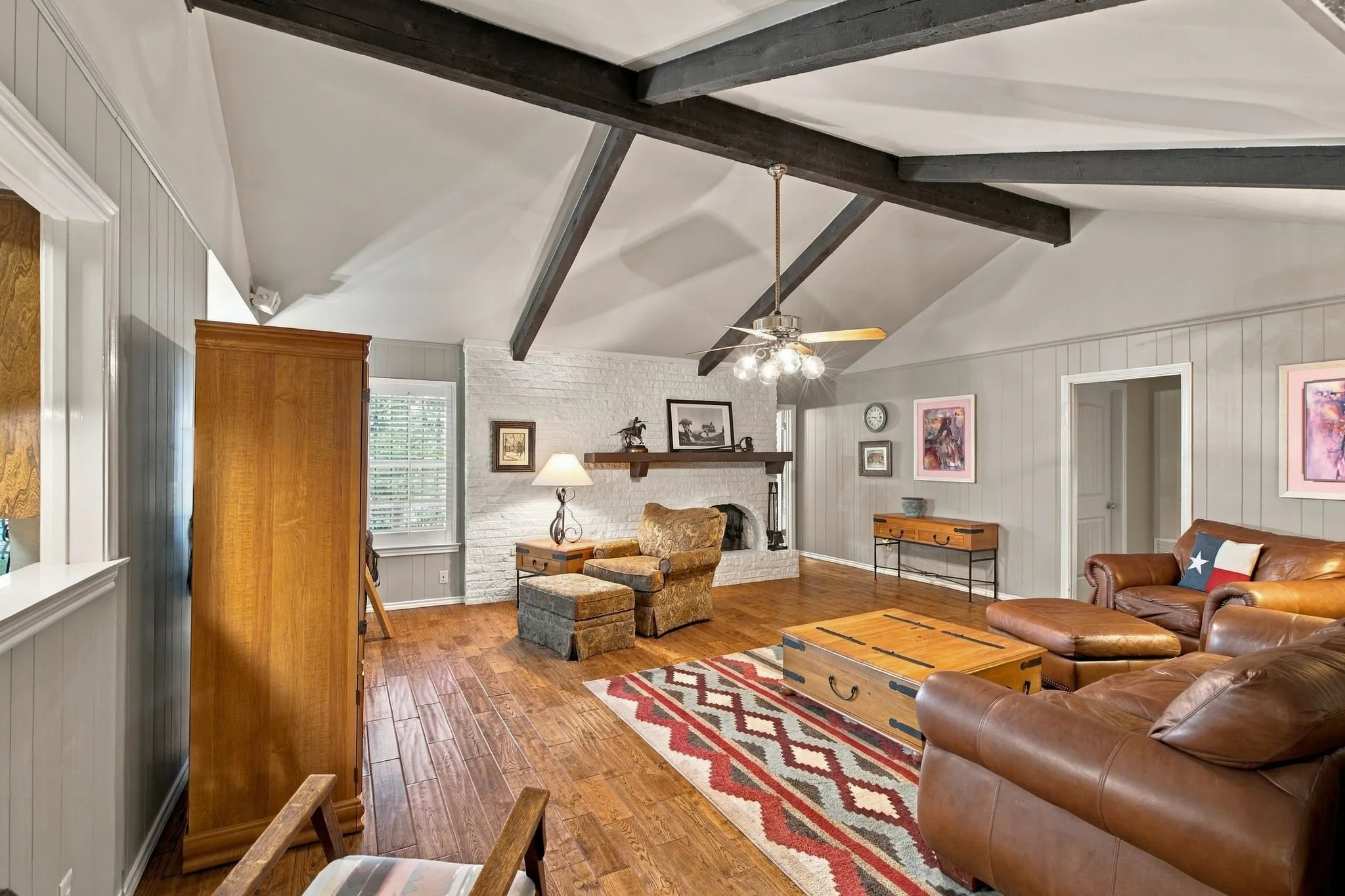Vaulted ceiling with exposed beams, wood-finish flooring, white brick fireplace, paneling accent wall, and a ceiling fan with integrated lighting