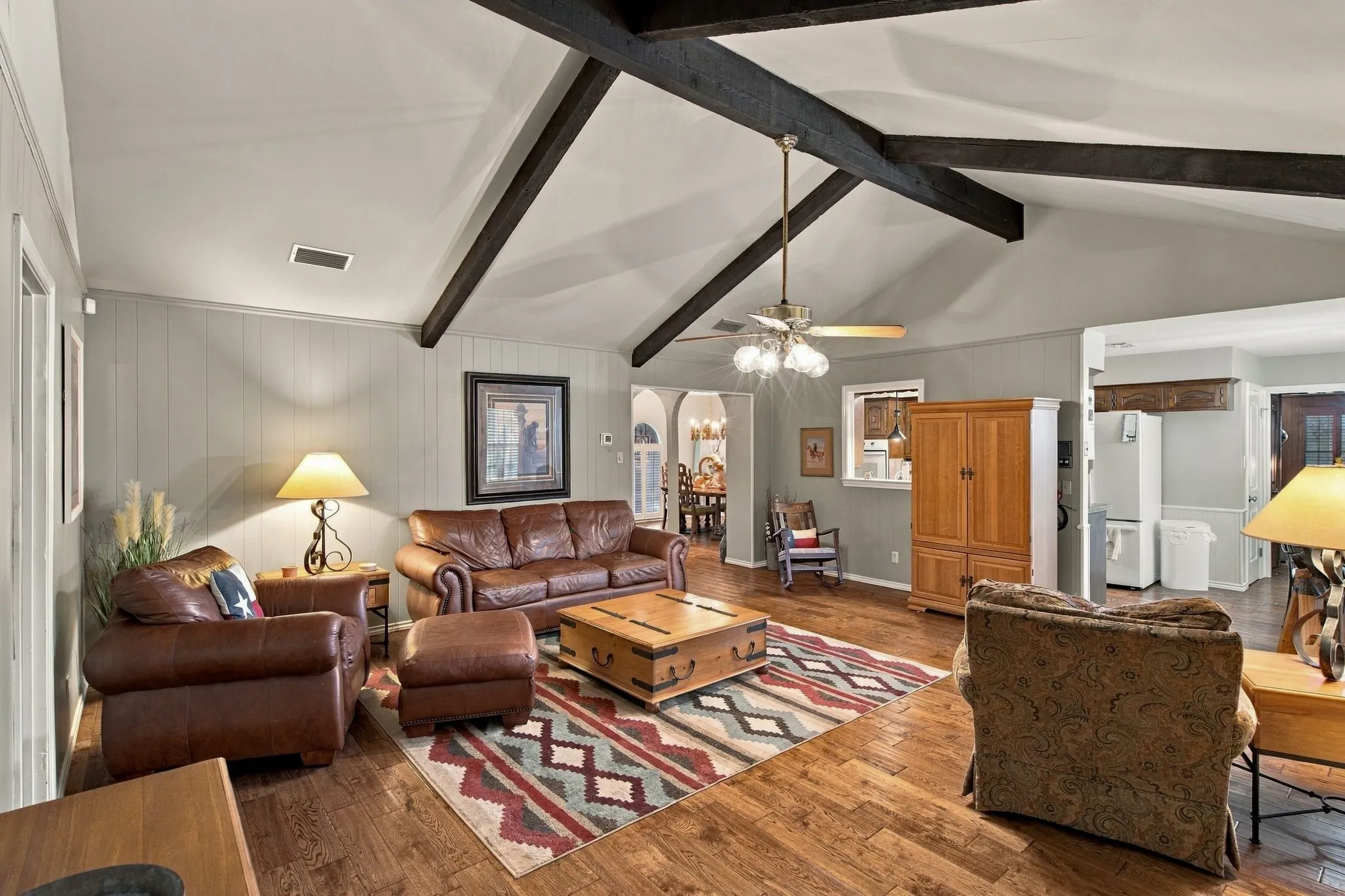 Vaulted ceiling with exposed beams, wood-finish flooring, paneled wall detailing, and a ceiling fan with integrated lighting