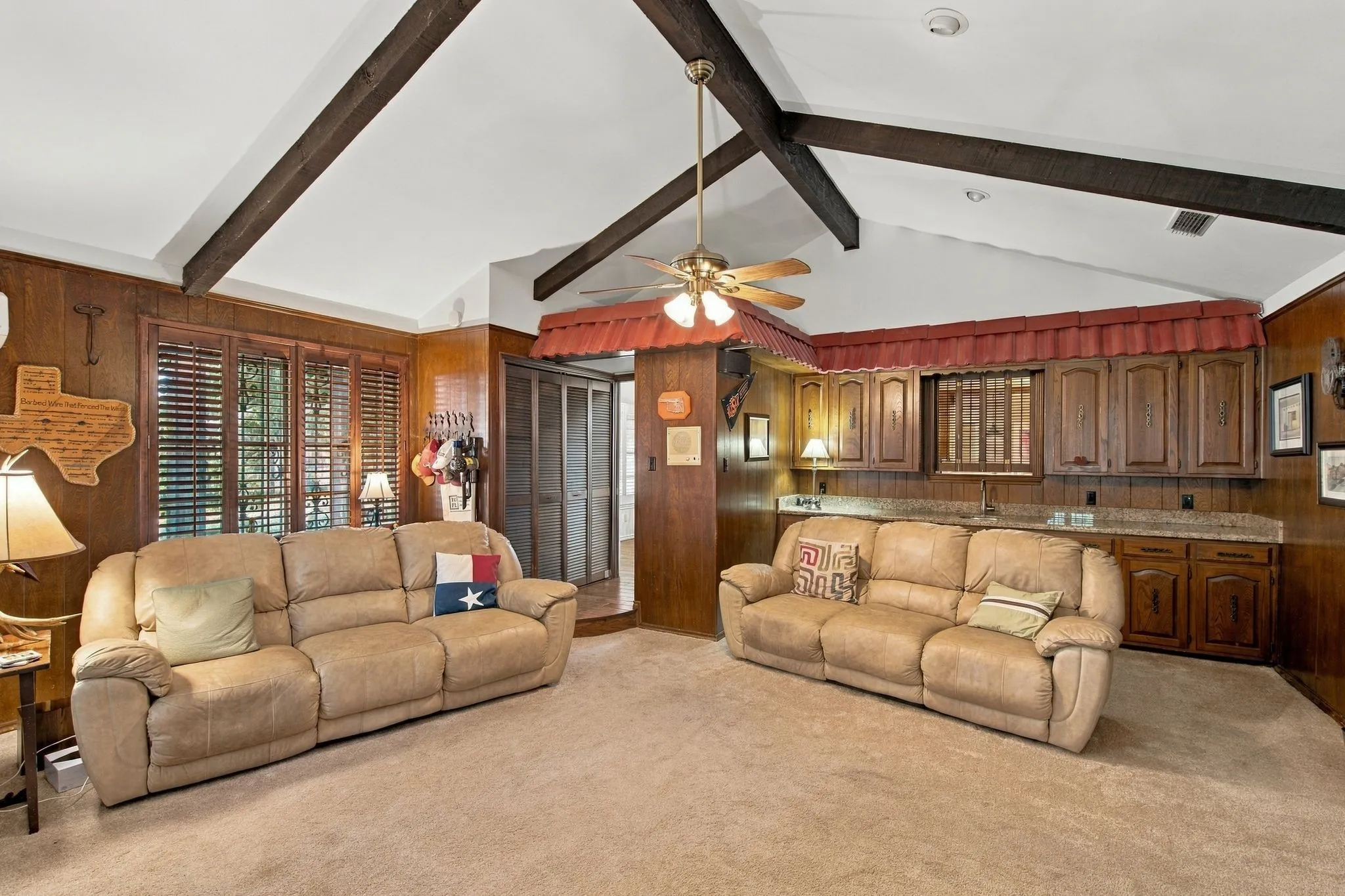 Vaulted ceiling with exposed beams, wood paneling, built-in wet bar with cabinet storage and stone-finish countertop, plantation shutters, and ceiling fan
