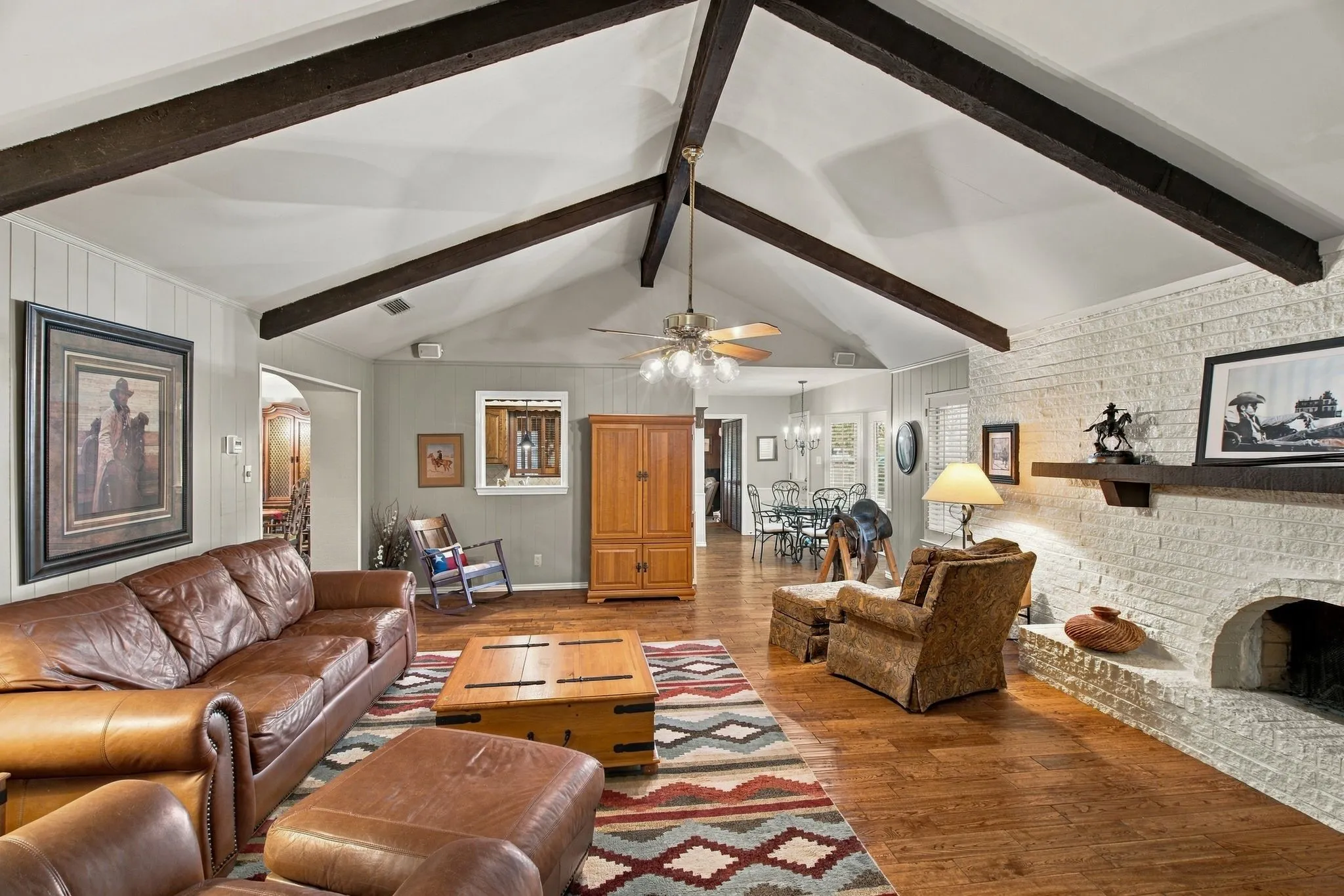 Vaulted ceiling with exposed wood beams, wood-finish flooring, white brick fireplace with hearth and wood mantel, ceiling fan with integrated lighting, and paneled wall finishes