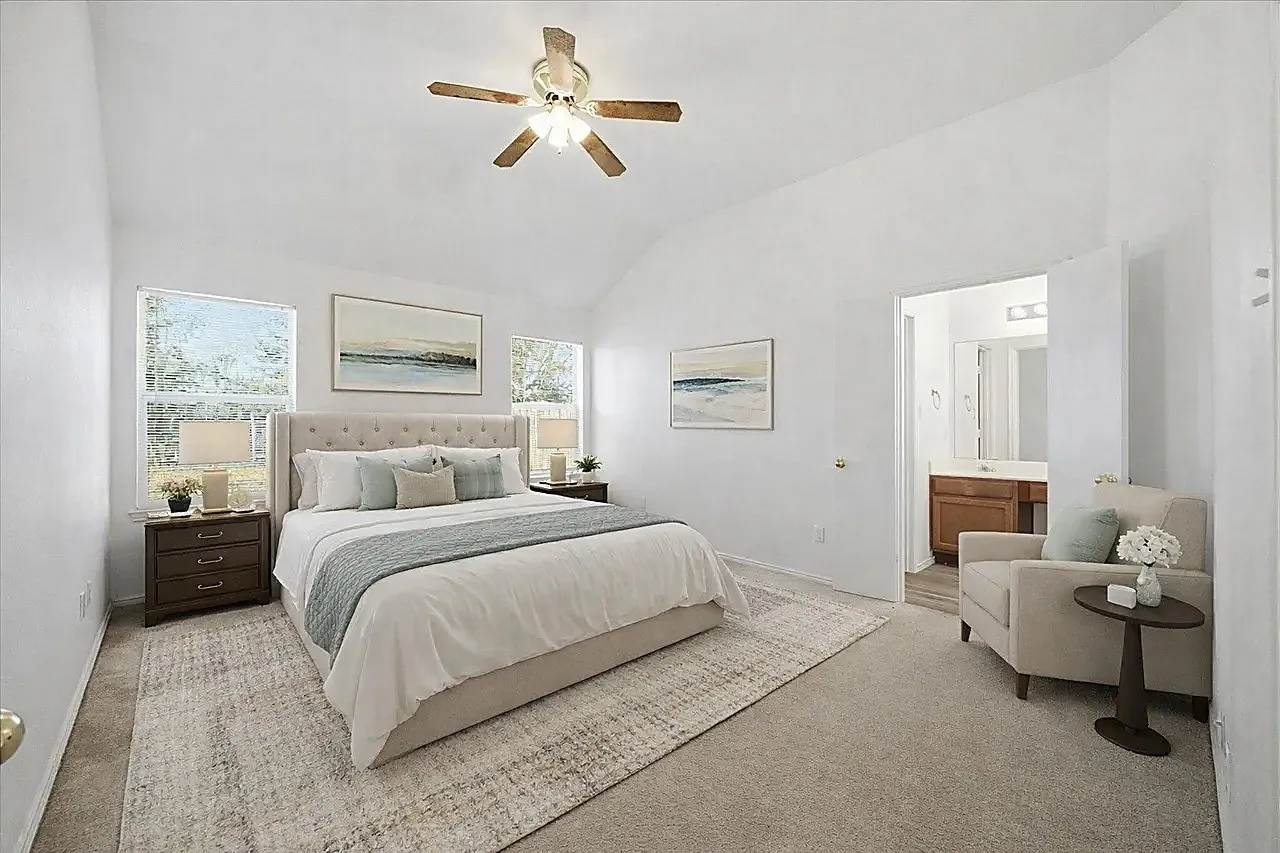 Vaulted ceiling with a ceiling fan, dual windows, carpeted flooring, and a visible ensuite bathroom with a wood-finish vanity