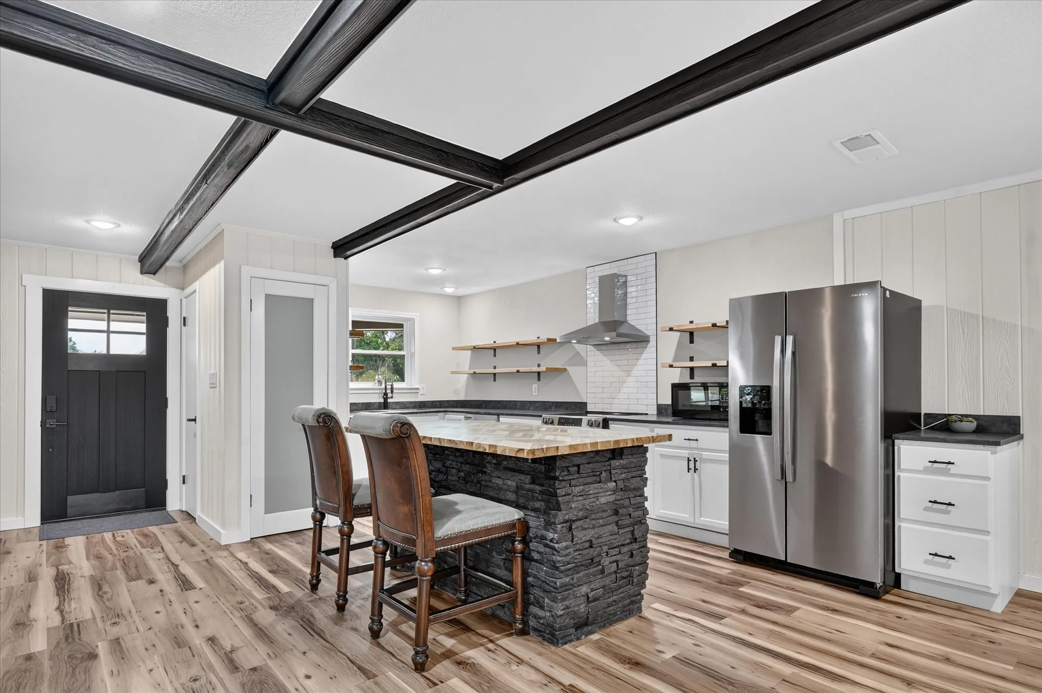 Open-concept kitchen with wood-finish flooring, exposed ceiling beams, a stone-clad island, white shaker cabinetry, and stainless steel appliances