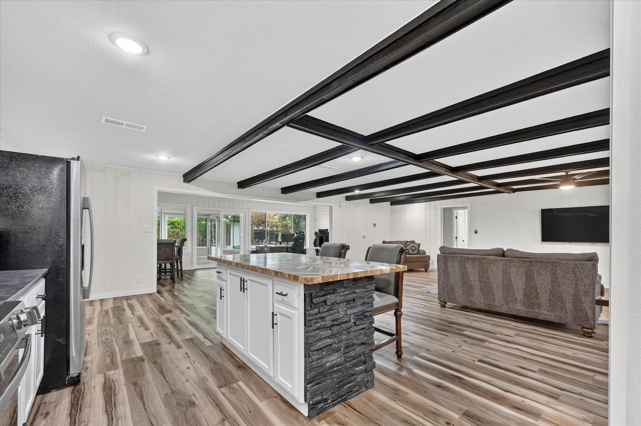 Open-concept kitchen and living area featuring a large island with a wood-finish top and gray stone base, white cabinetry with stainless steel appliances, wood-finish flooring throughout, exposed black beam ceiling details, and abundant natural light from multiple windows and glass doors