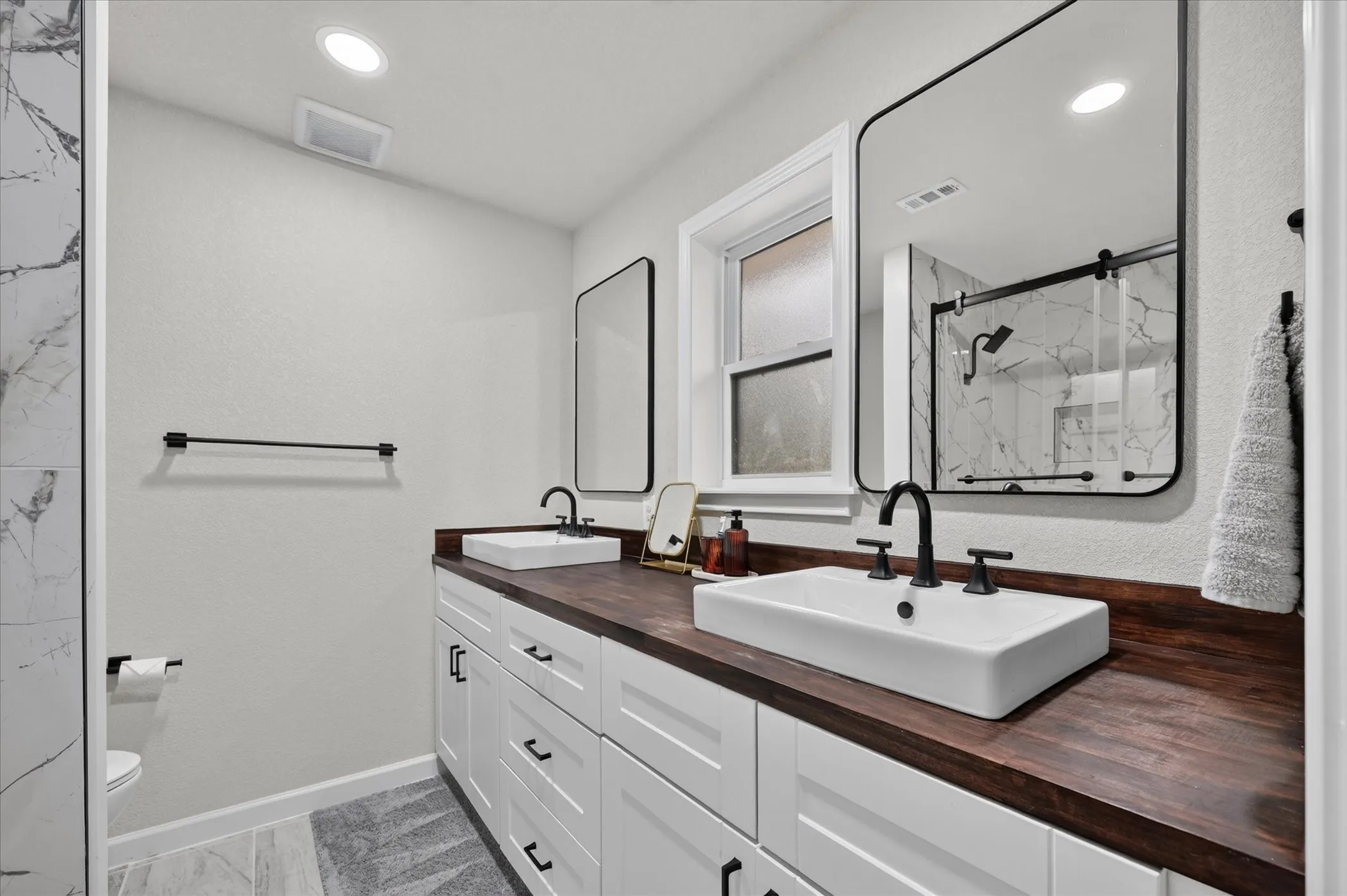 Dual vessel sinks with matte black fixtures, white shaker-style cabinetry, rich wood-finish countertops, and rectangular framed mirrors