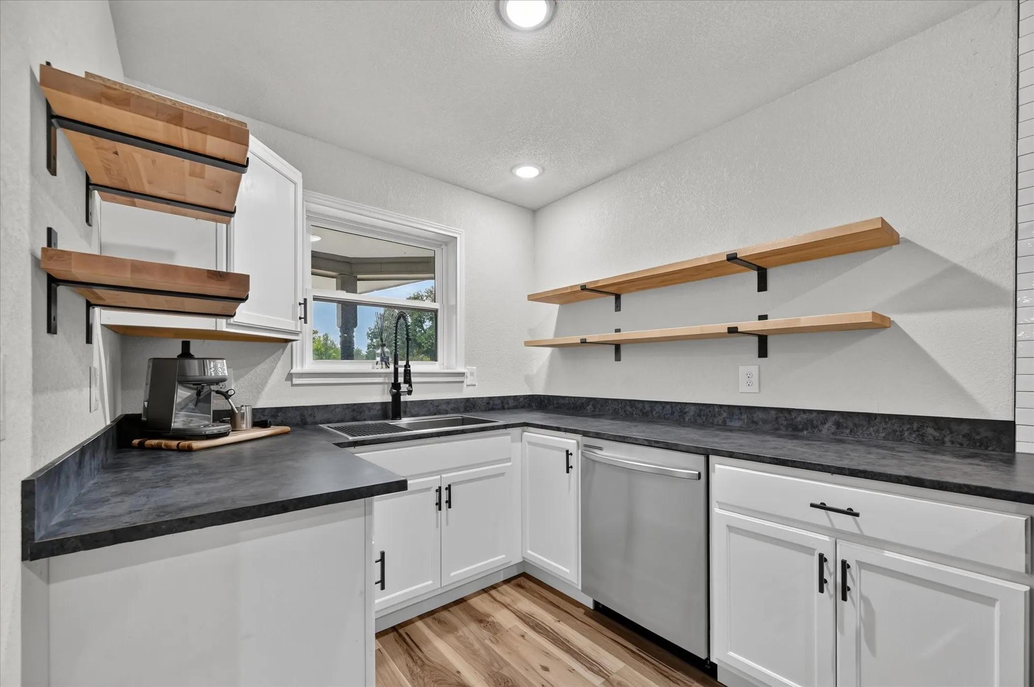 White cabinetry with matte black hardware, dark countertops, wood-finish flooring, stainless steel dishwasher, and open wood shelving