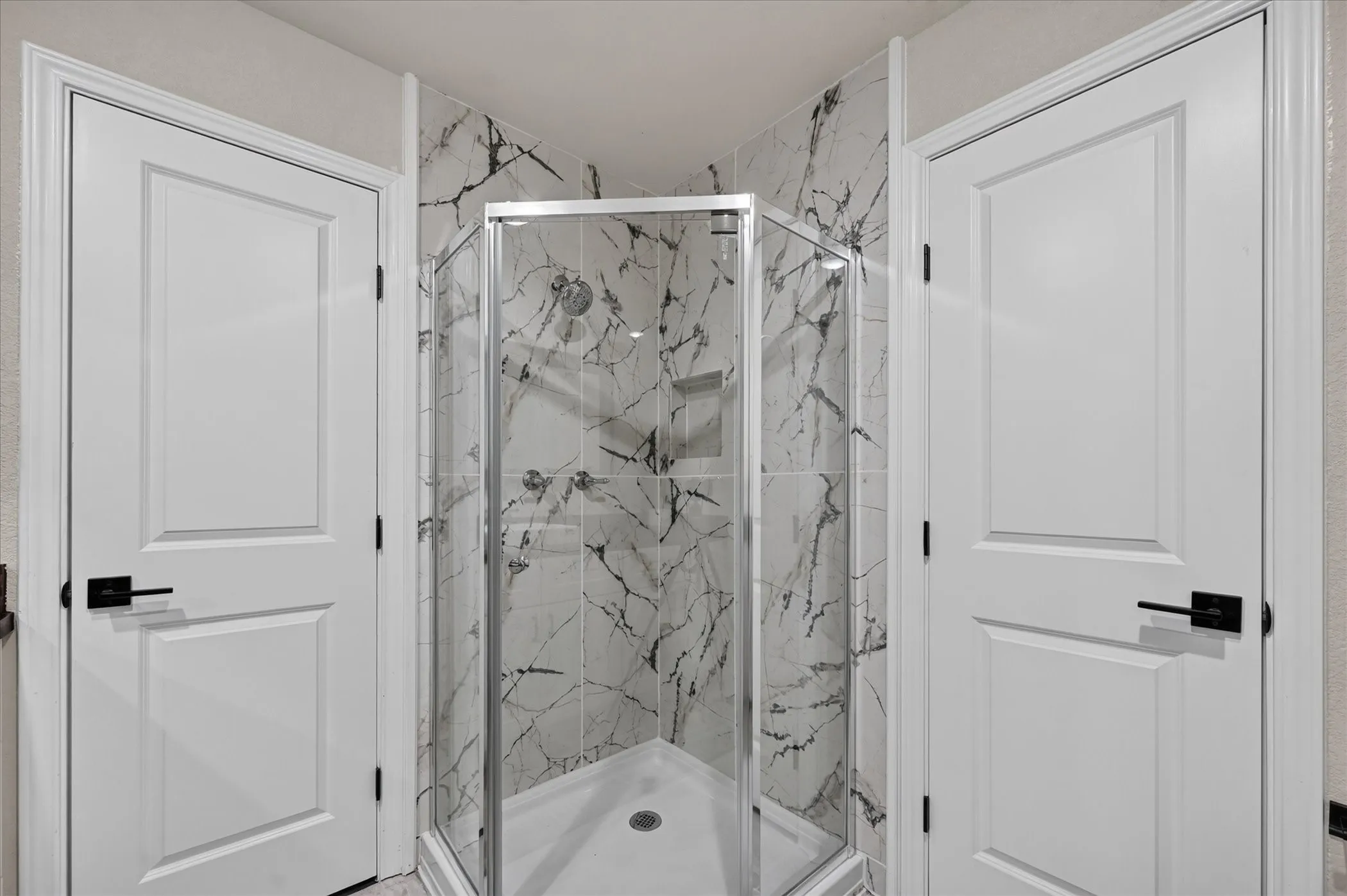 Corner shower with glass enclosure, white tile surround with grey veining, built-in shower niche, and two white panel doors with matte black hardware