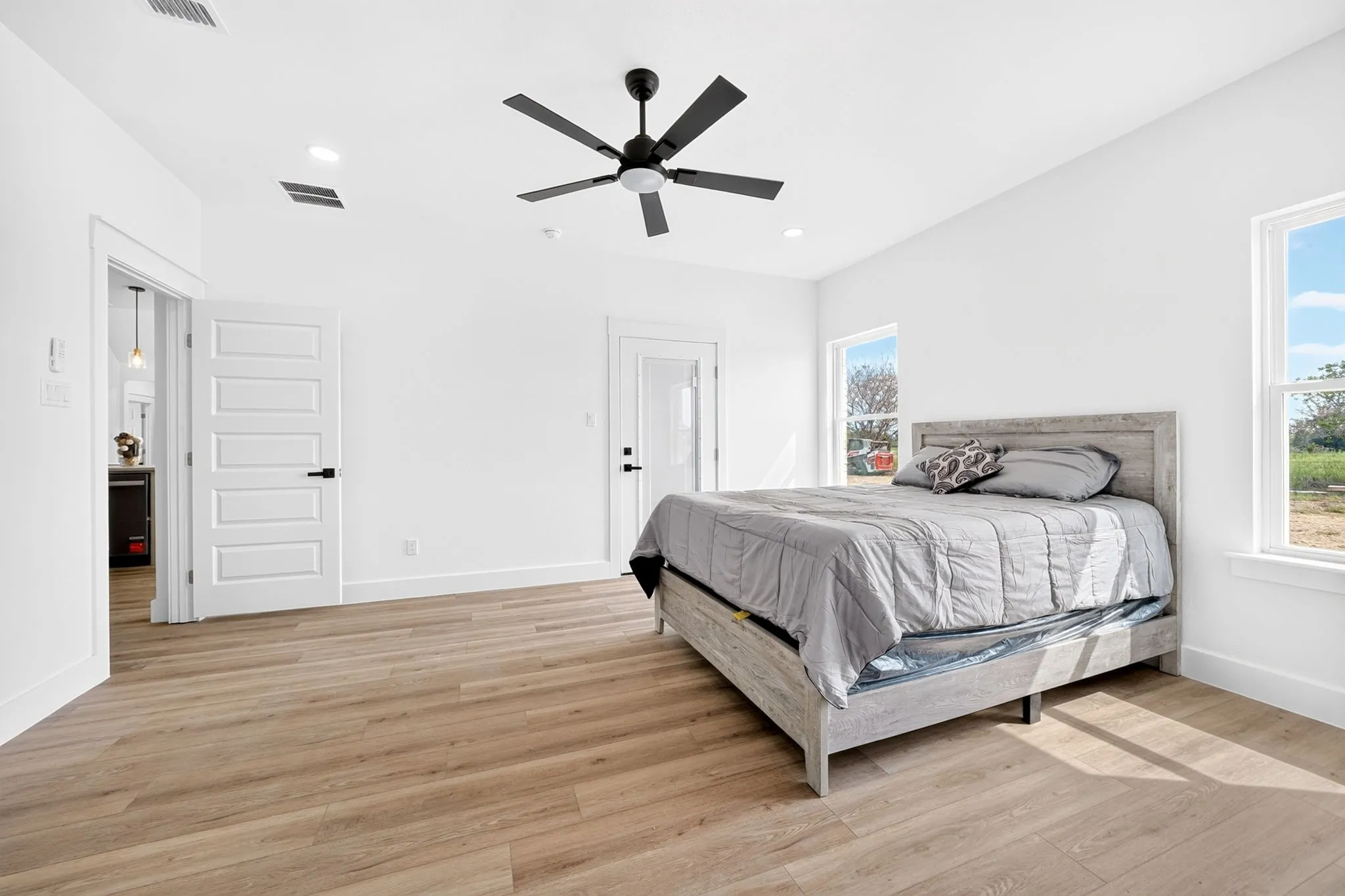 Recessed ceiling lighting, wood-finish flooring, two windows with white trim, a ceiling fan with a matte black finish, and a white paneled door with a matte black handle