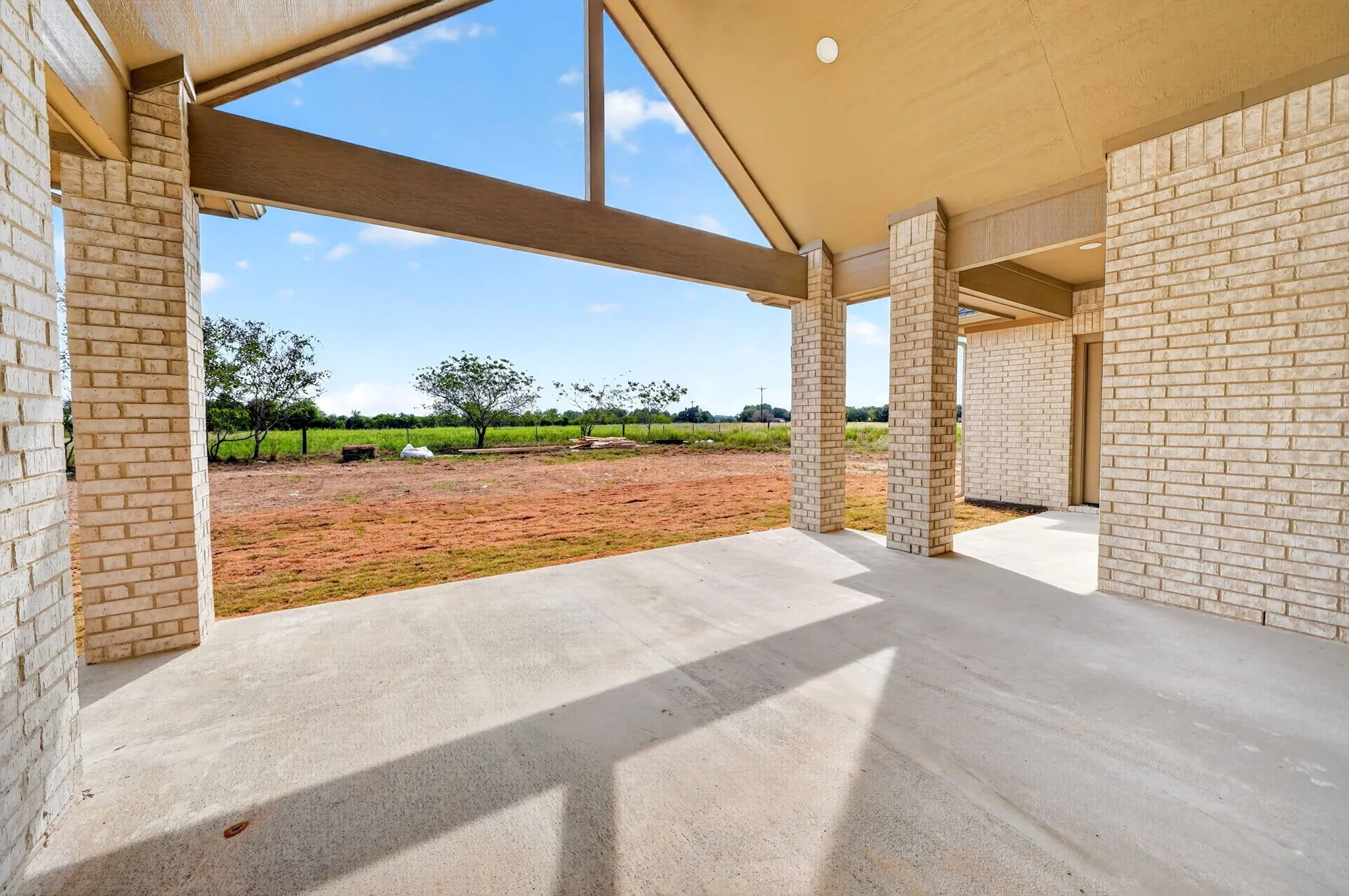 Expansive covered concrete patio with brick pillars, exposed beam ceiling, and a broad open-sky backdrop