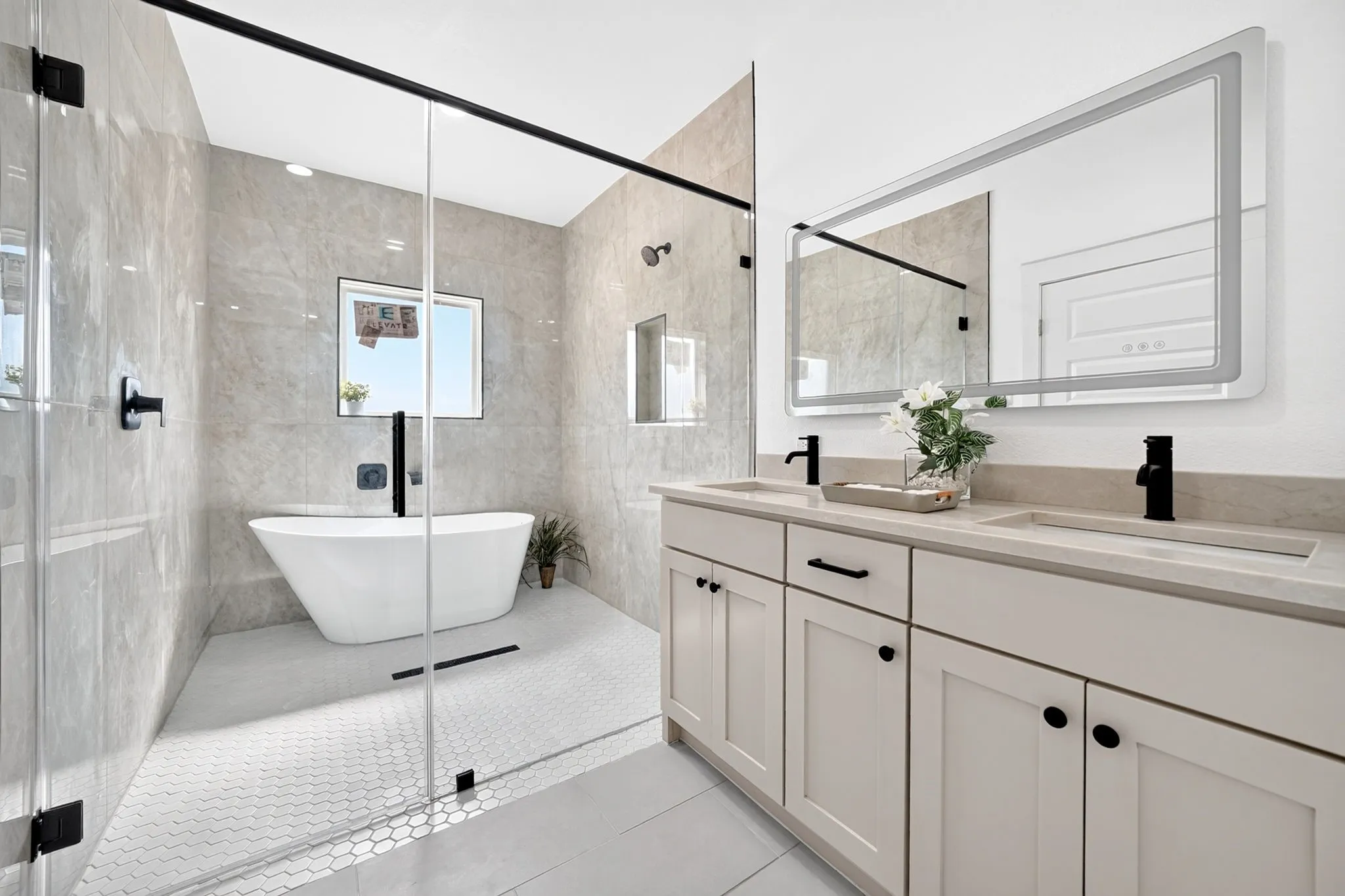 Dual vanity with undermount sinks and matte black fixtures, an illuminated mirror, a freestanding soaking tub, a walk-in shower with a glass enclosure, and large format wall tiles
