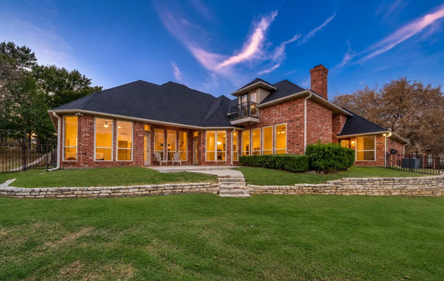 Stunning twilight view showcases a grand brick home, glowing windows, stone landscaping, and expansive backyard lawn