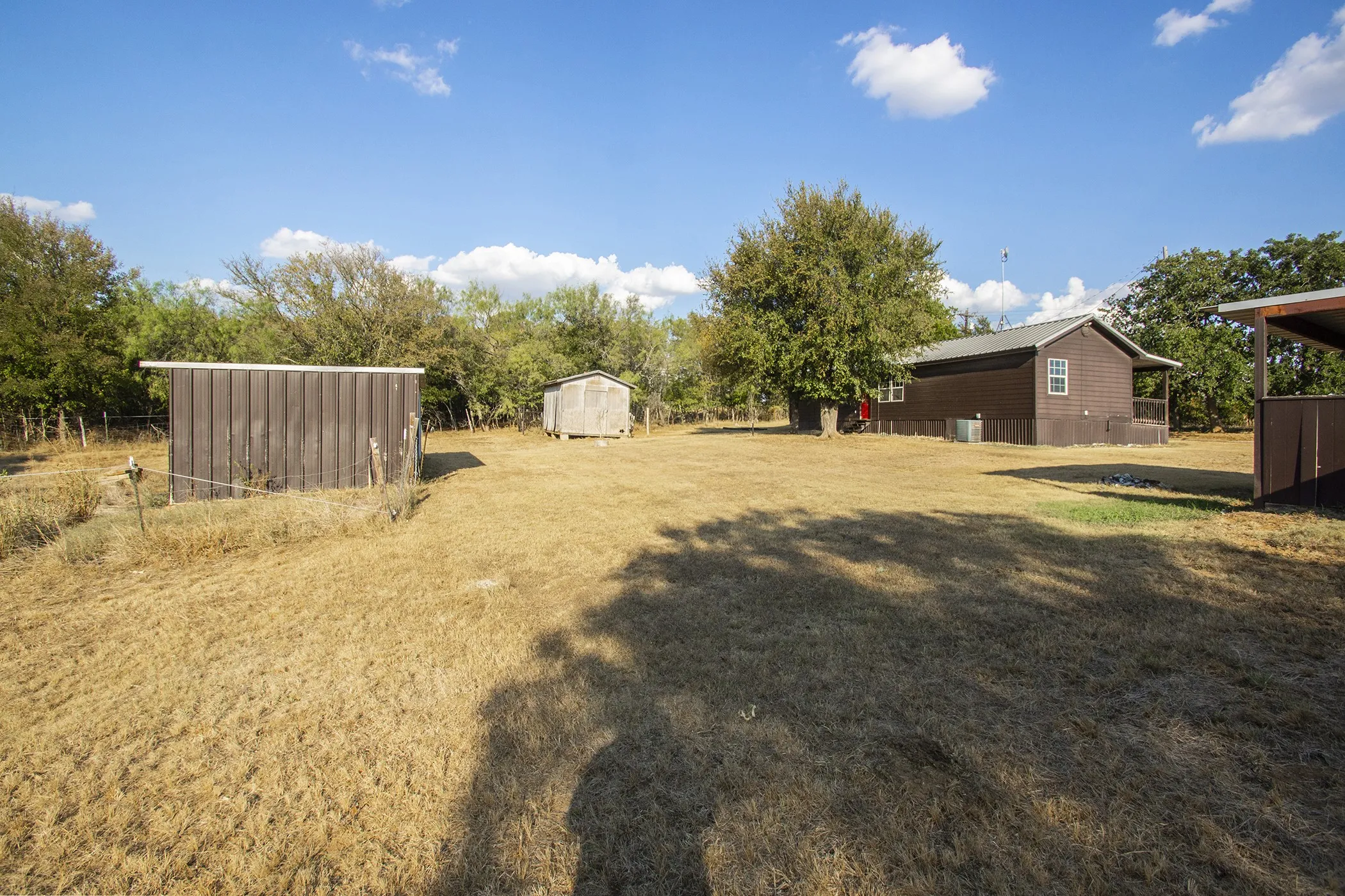 Expansive outdoor space featuring a brown single-story structure, a second auxiliary structure, and mature trees
