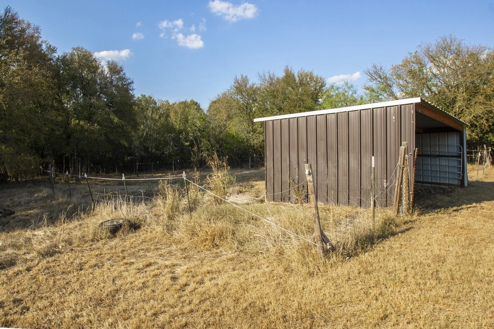 Metal-sided shed with a single open bay, a corrugated roof, and a partial wire fence enclosure