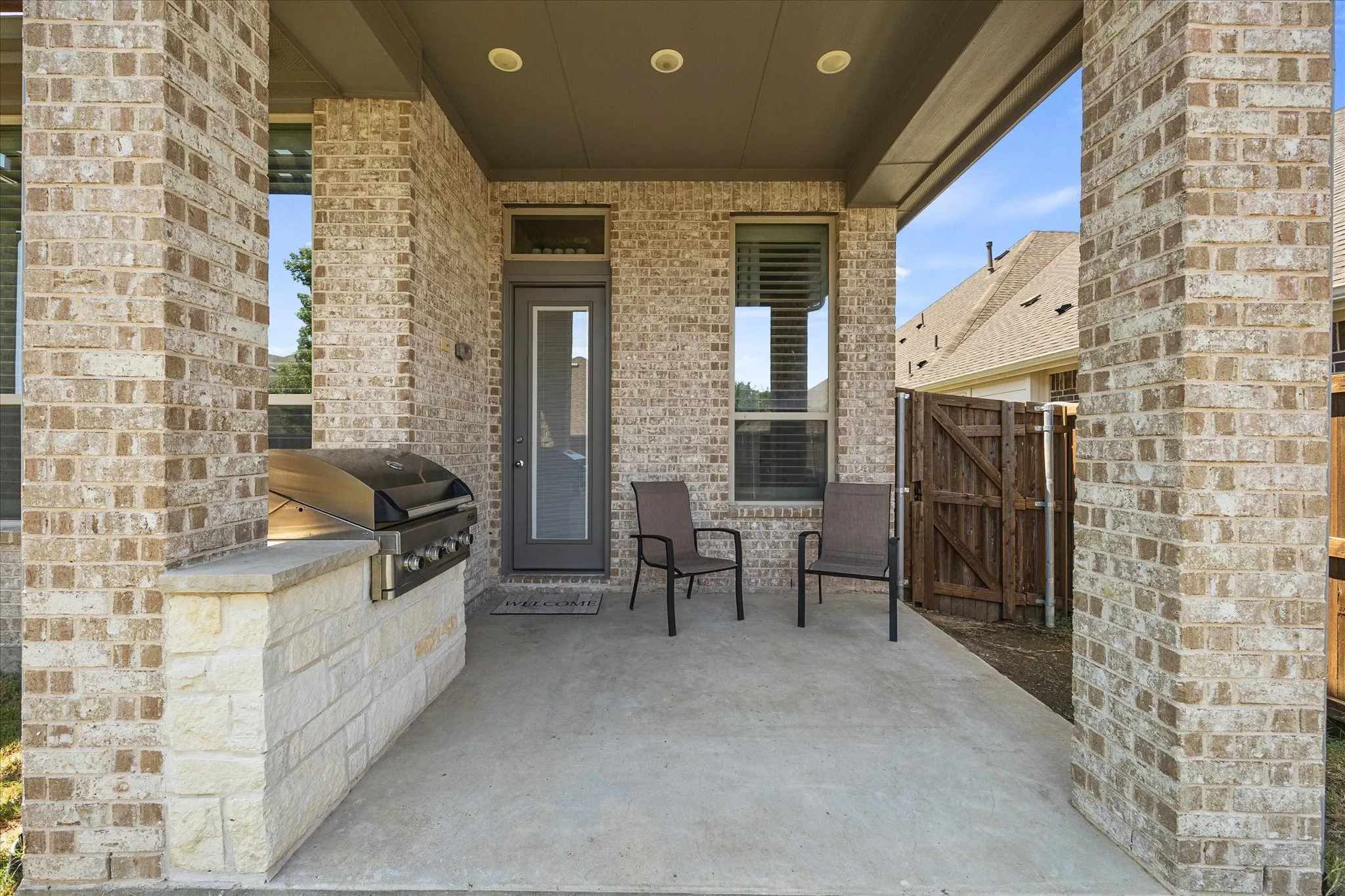 Covered patio featuring a built-in outdoor grill with a stone-finish base, brick columns, and a privacy fence