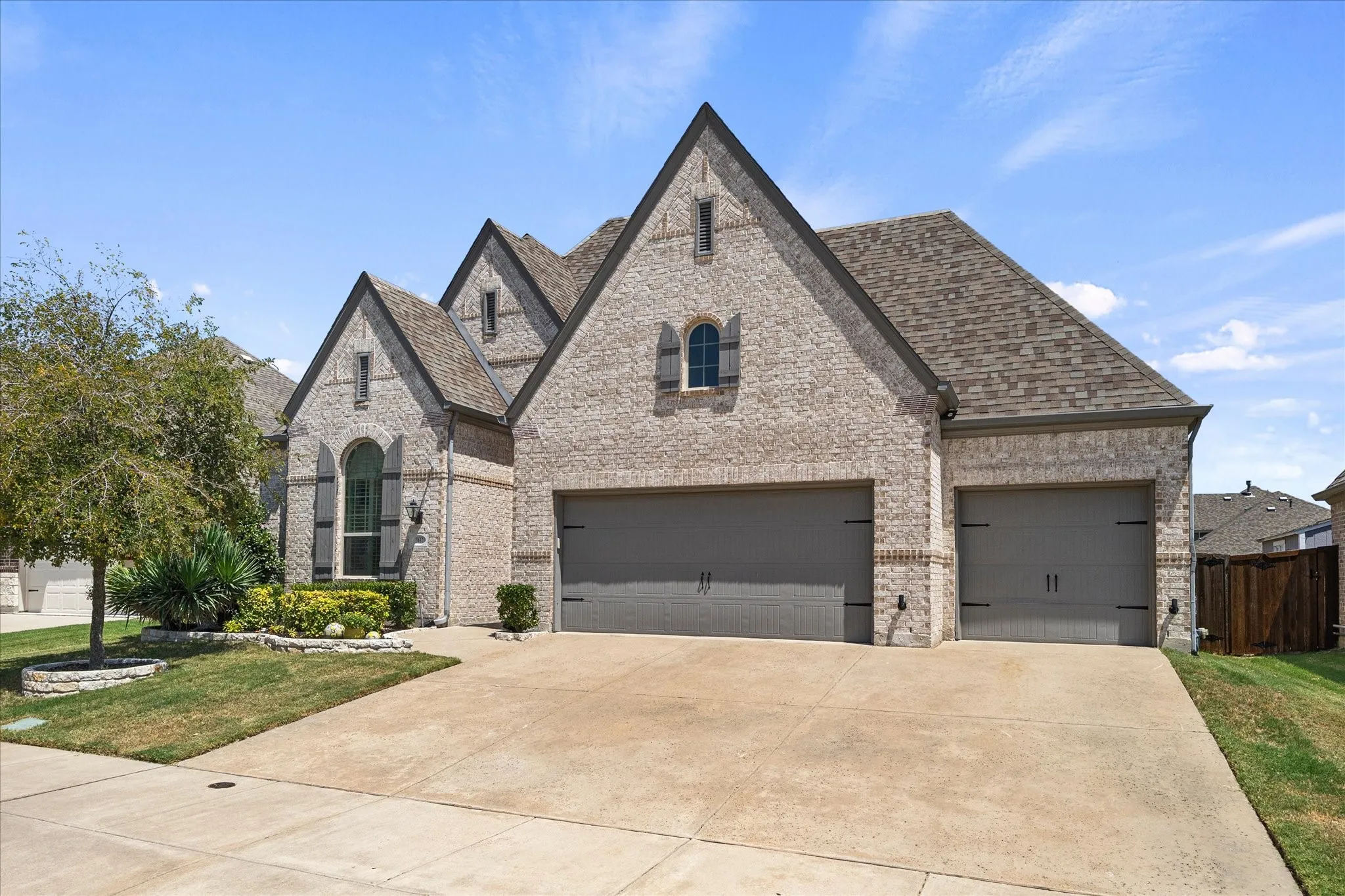 Brick exterior with a multi-gabled roofline, arched window with shutters, and a concrete driveway leading to two garage doors