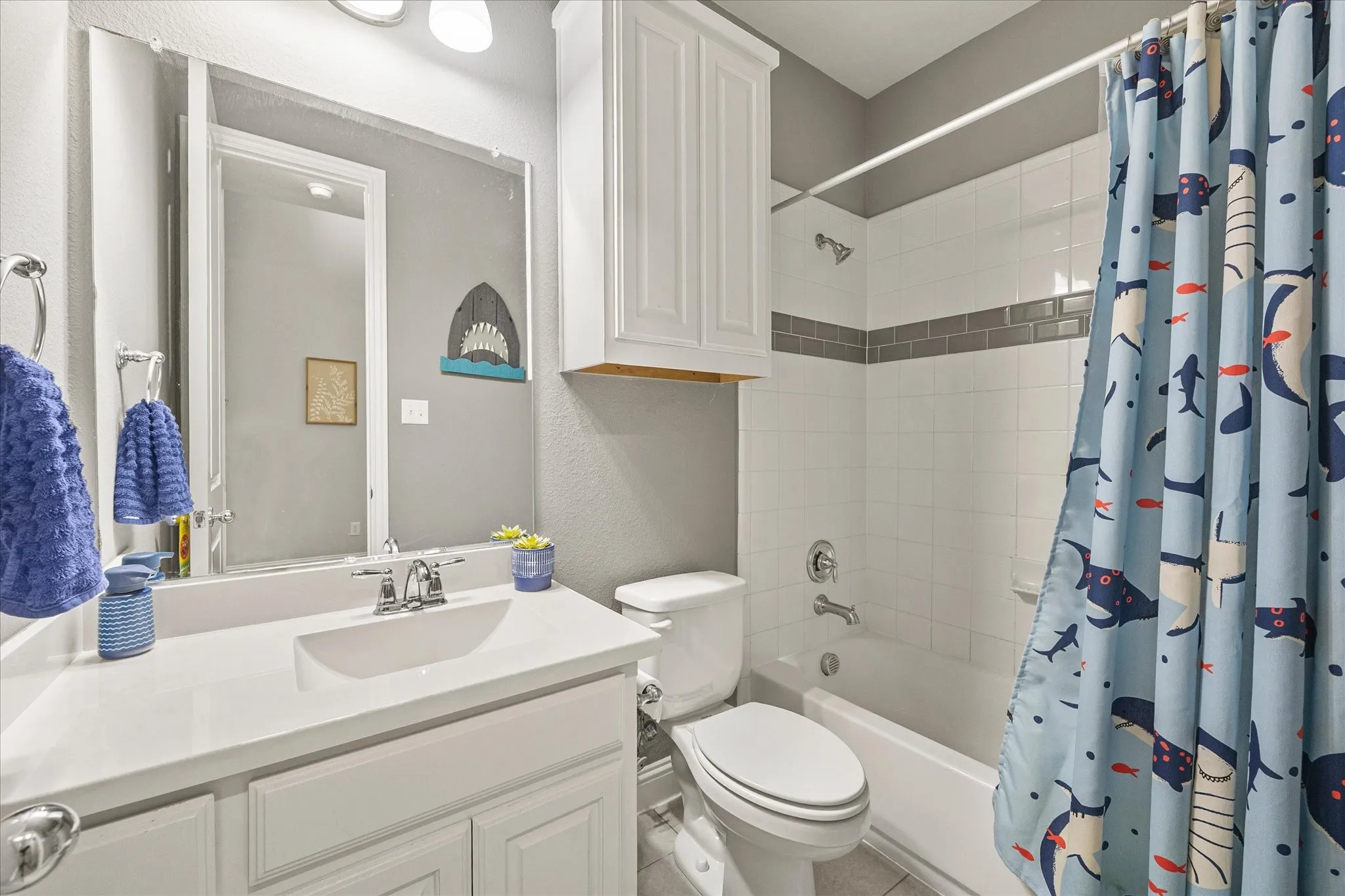 Vanity with integrated sink and chrome faucet, white cabinetry, overhead storage, white tiled shower surround with accent band, and built-in bathtub