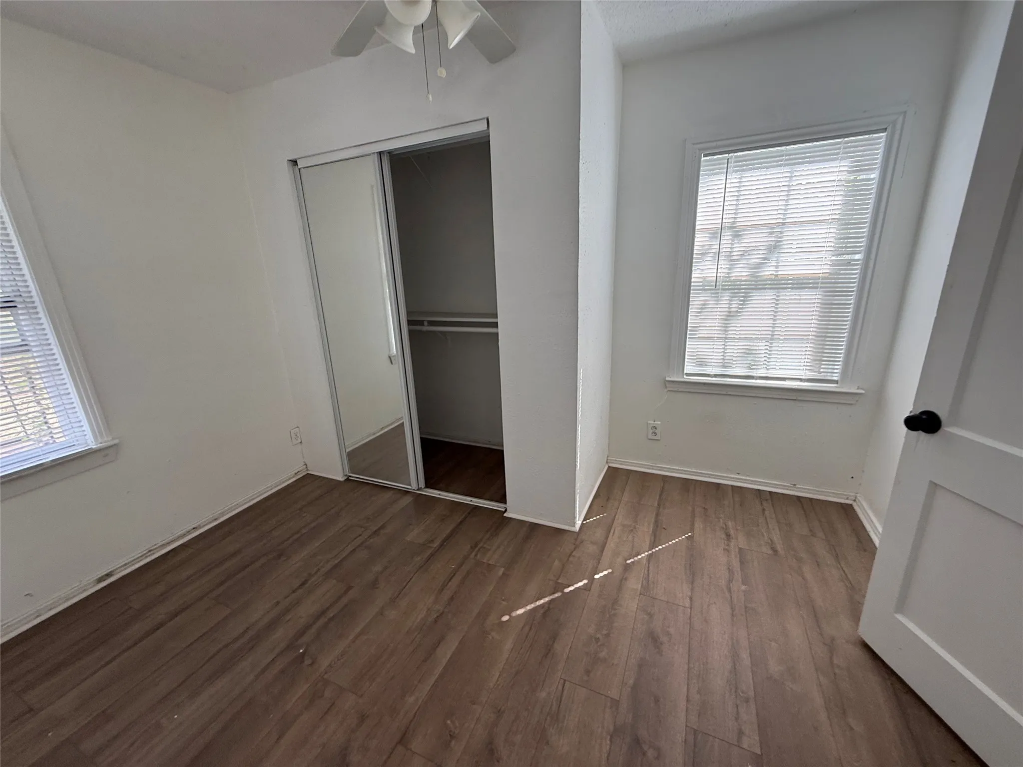 Wood-finish flooring throughout, white walls, two windows with blinds, mirrored sliding closet doors, and a ceiling fan