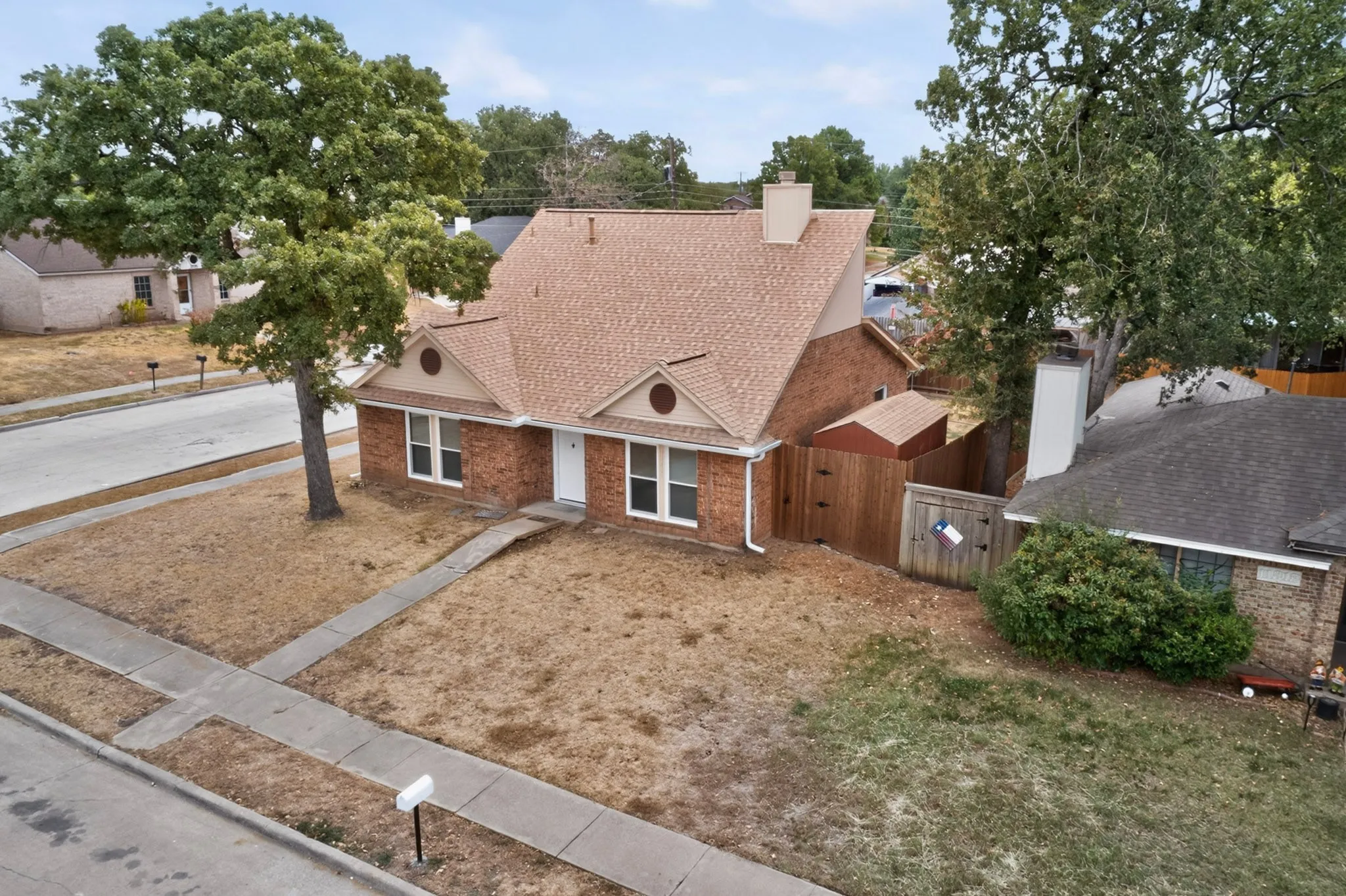 Brick exterior residence featuring a brown shingle roof, mature trees, and a wooden fence
