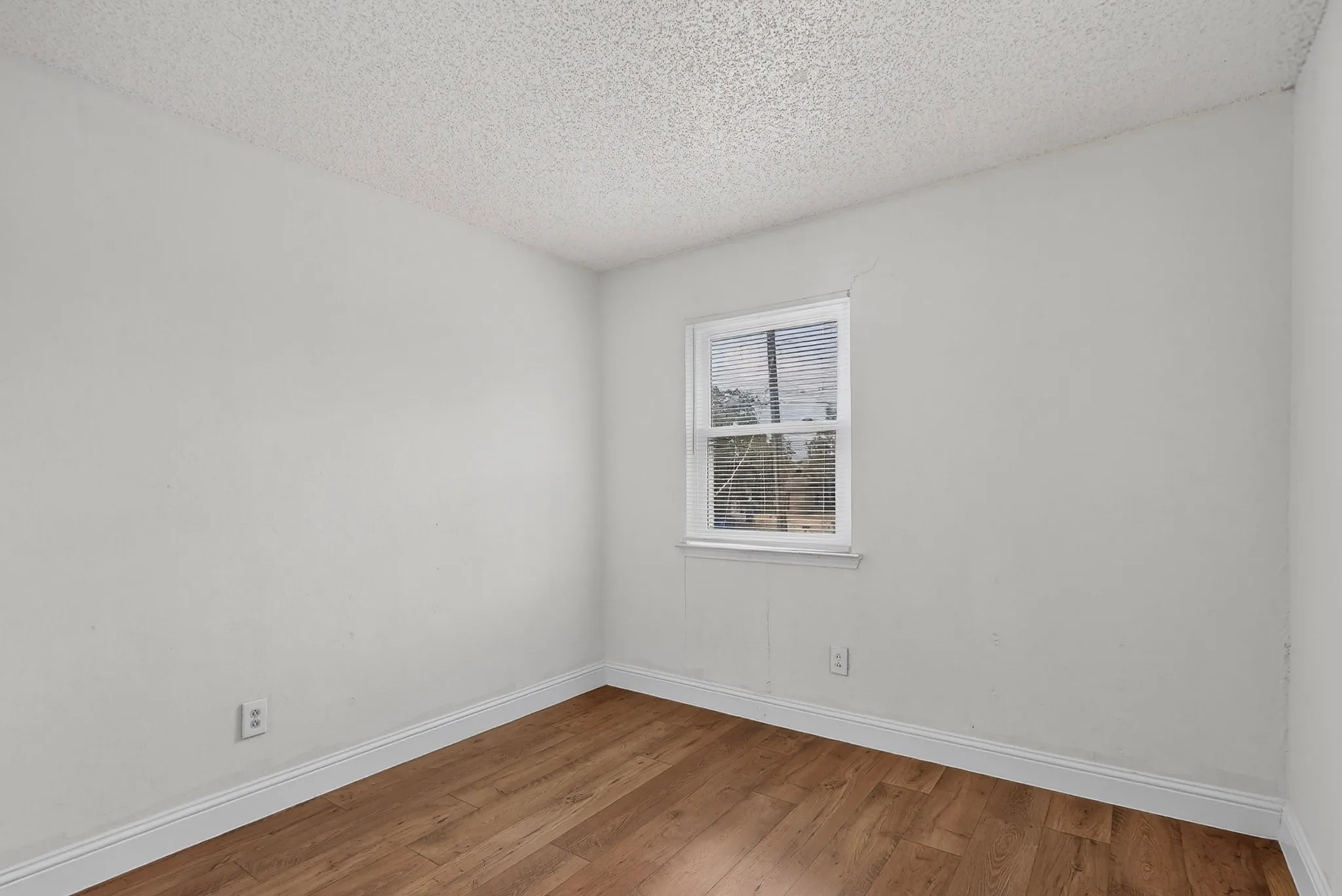 Wood-finish flooring with white trim, a single window with blinds, and white painted walls