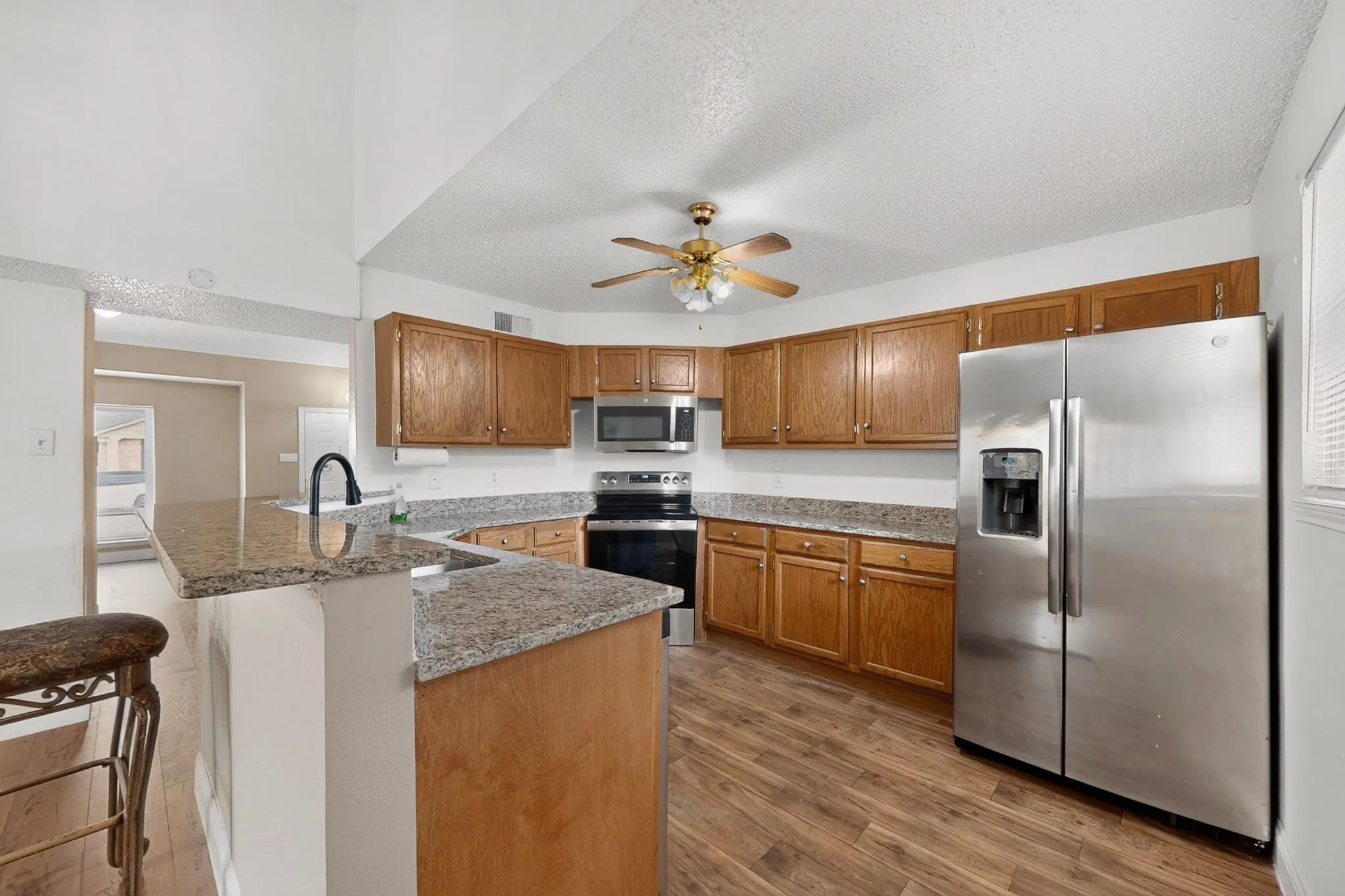 Kitchen featuring wood-finish flooring, L-shaped peninsula with stone-look countertop, wood cabinetry, built-in stainless steel appliances, and ceiling fan with lighting