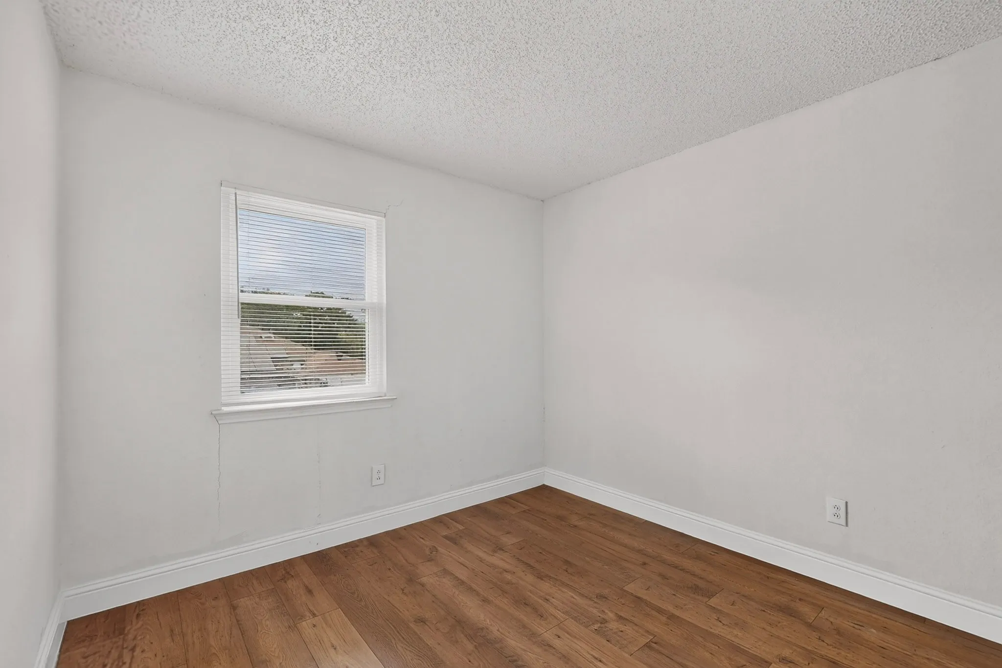 Wood-finish flooring with white baseboards and a window with blinds