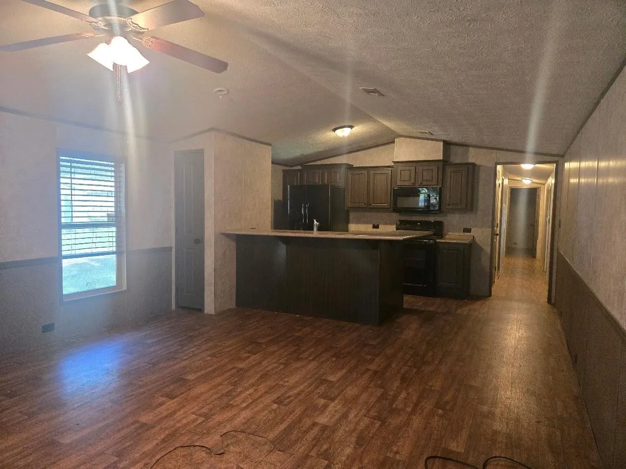 Open-concept living space featuring wood-finish flooring, a ceiling fan with integrated lighting, and a kitchen with dark cabinetry and black appliances