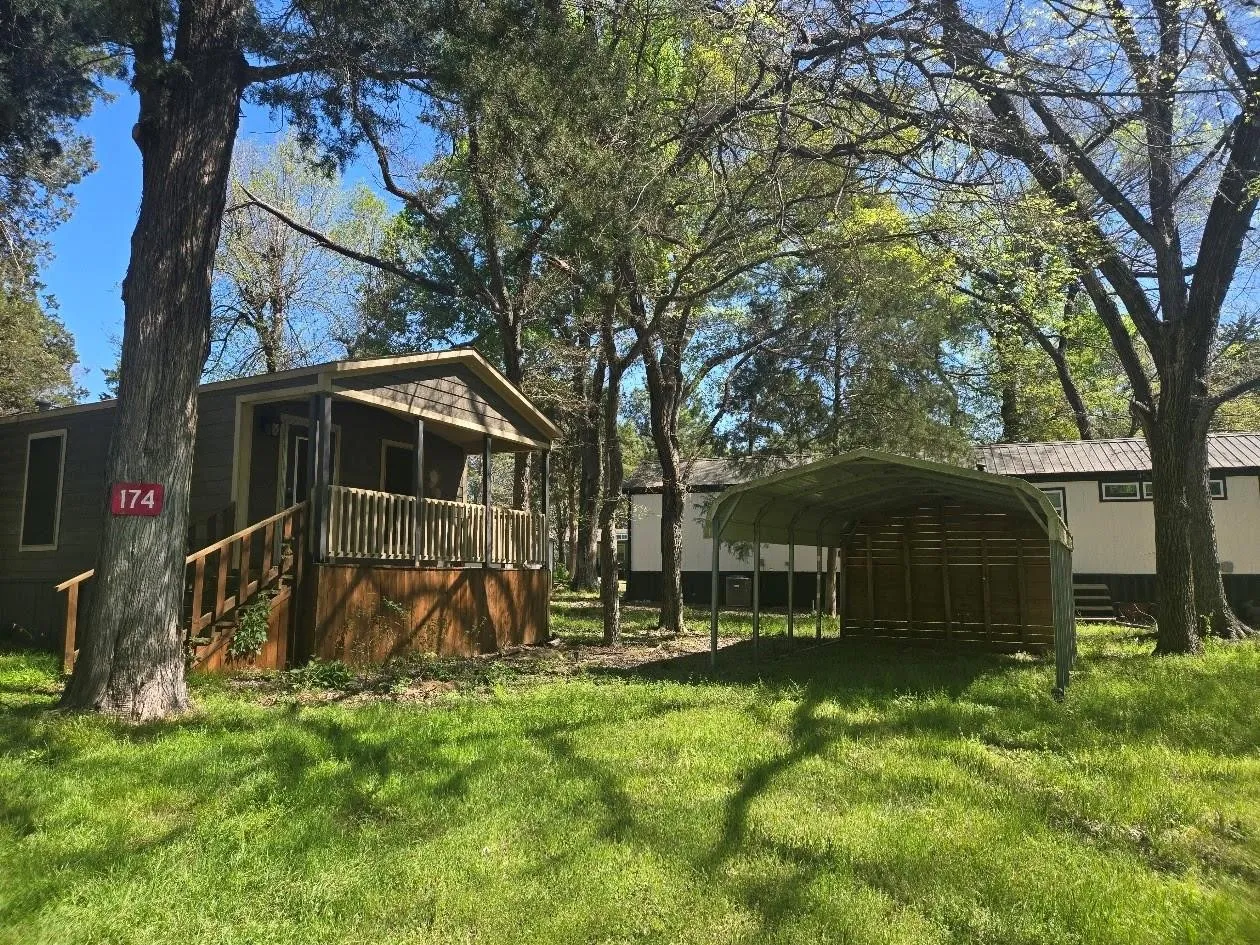 Covered porch entry, wood railing, wood-finish siding, metal carport structure, and verdant surroundings