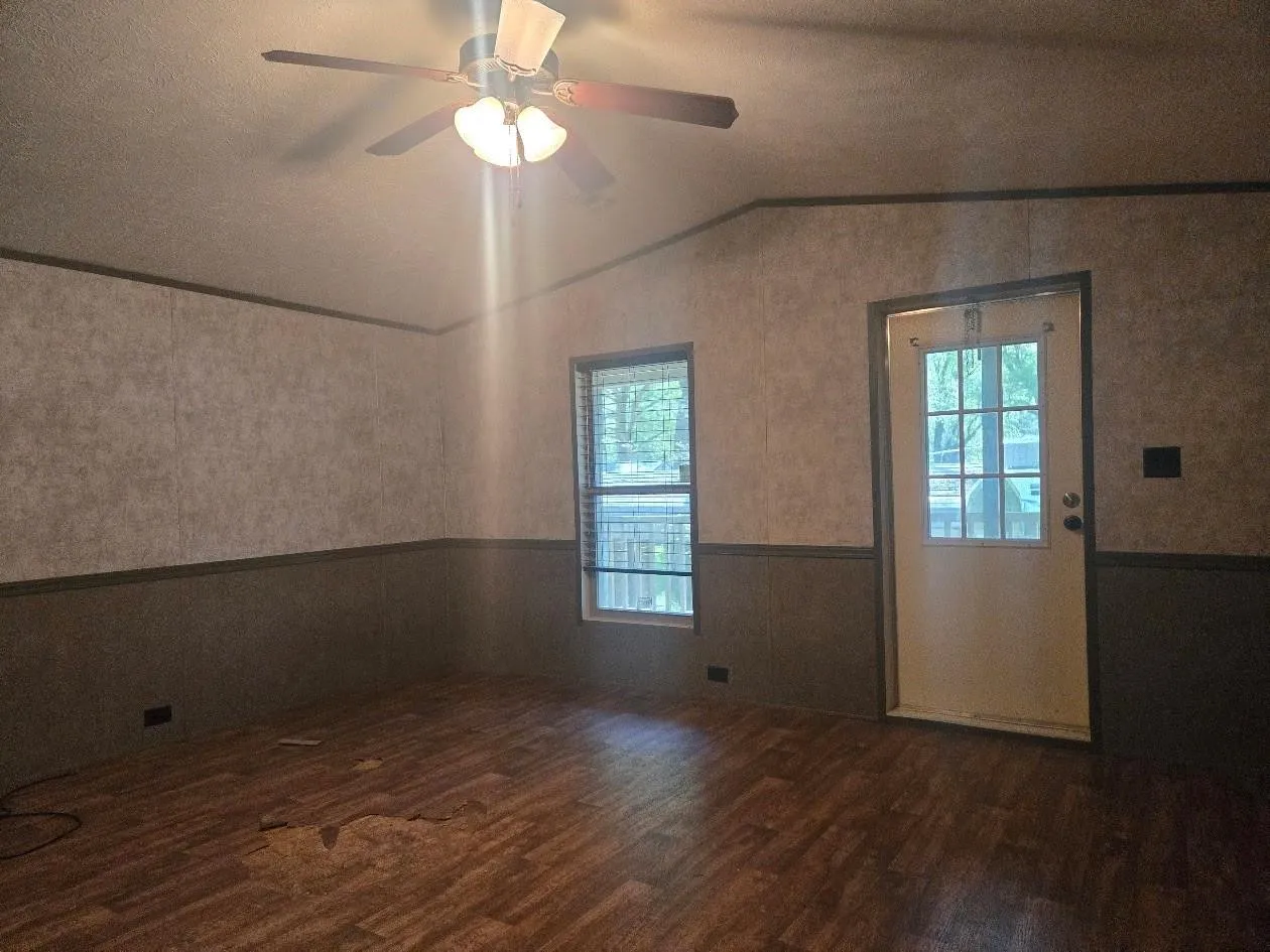 Wood-finish flooring, ceiling fan with integrated lighting, paneled walls, glass-paneled entry door, and a window with blinds