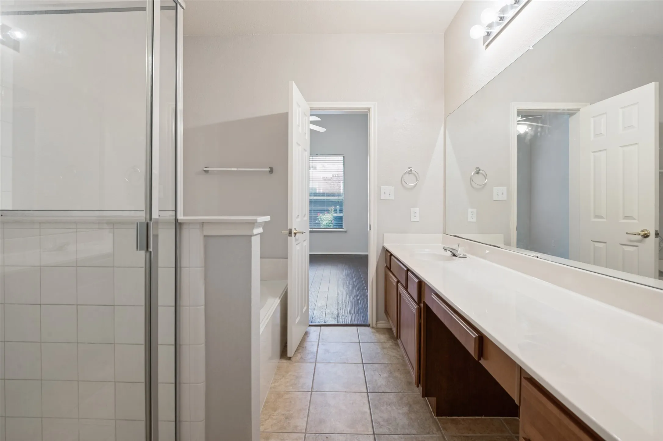 Glass-enclosed shower, built-in bathtub, extended vanity with dual sinks, wall-mounted mirror, and tile flooring