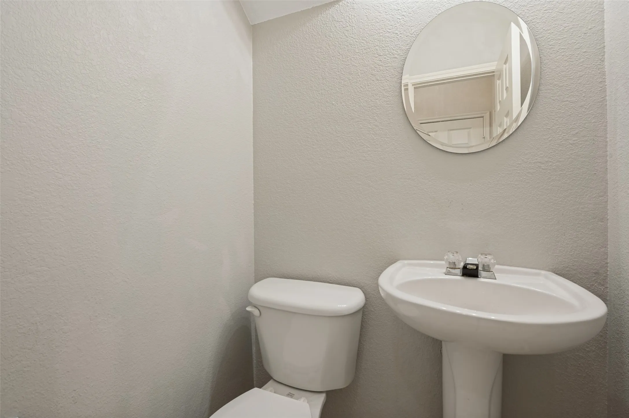 Pedestal sink with a two-handle faucet, circular wall mirror, and a white toilet