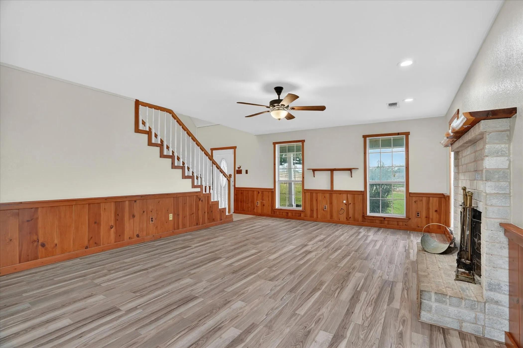 Wood-finish flooring, wood panel wainscoting, white painted walls, brick fireplace with wood mantel, and a ceiling fan with light fixture