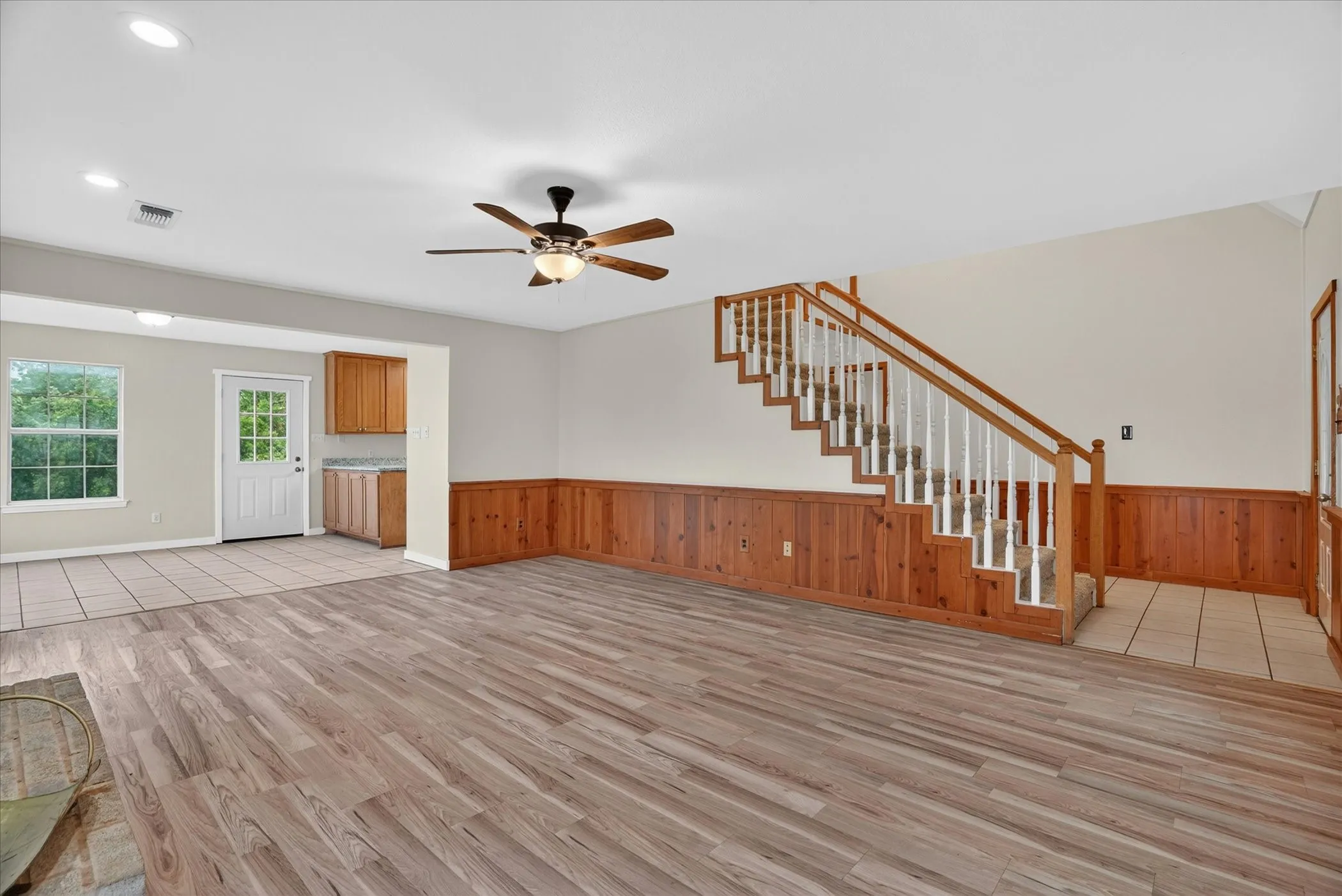 Spacious living area featuring wood-finish flooring, a ceiling fan with integrated lighting, and natural wood wainscoting