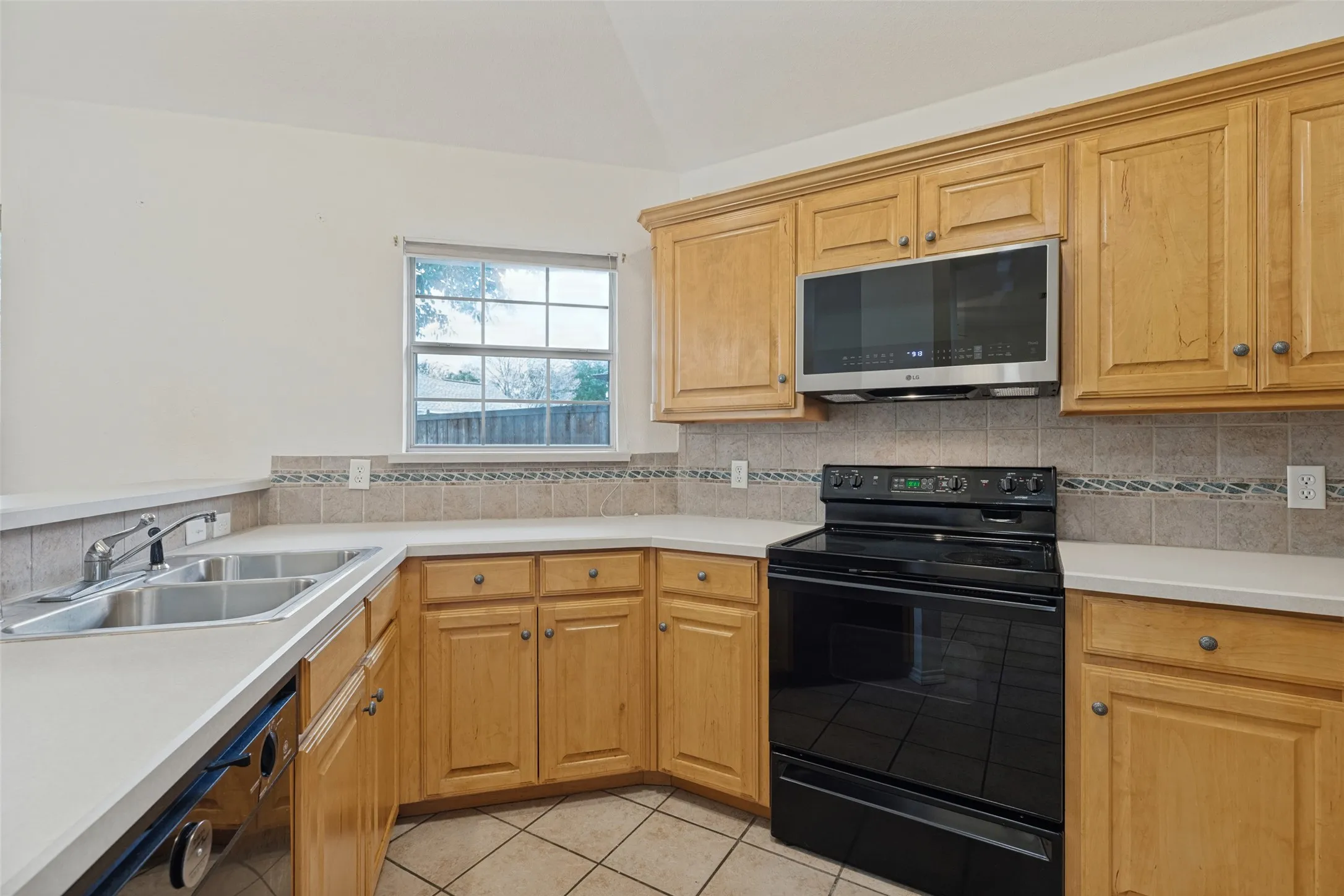 Light wood cabinetry, tile backsplash with decorative accent, built-in microwave, dual basin stainless steel sink, and tile flooring